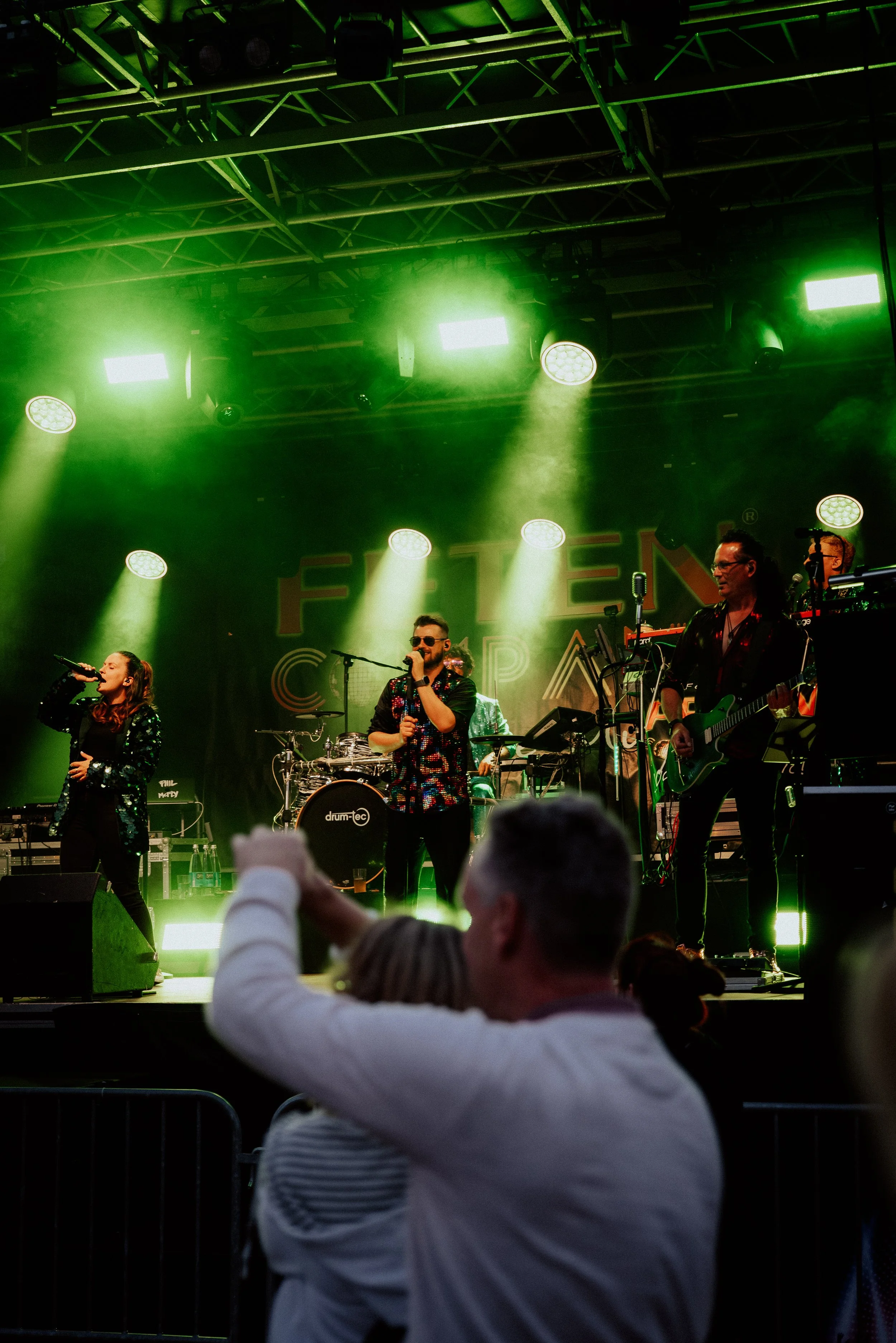 Auf der Bühne einer Musikshow in grüner Beleuchtung performen Sänger und Musiker. Im Vordergrund schaut ein Zuschauer mit weißem Pullover, der ein Mobiltelefon hebt, um den Auftritt aufzunehmen.