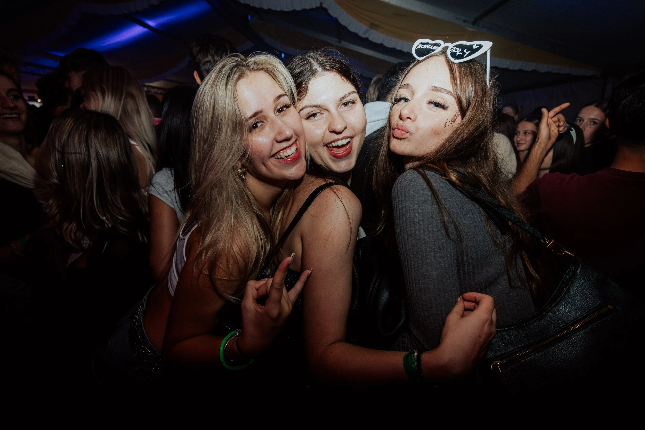Drei junge Frauen feiern auf einer Party, lachen und posieren für die Kamera. Eine trägt eine Brille mit Herzformen und die anderen lächeln fröhlich.