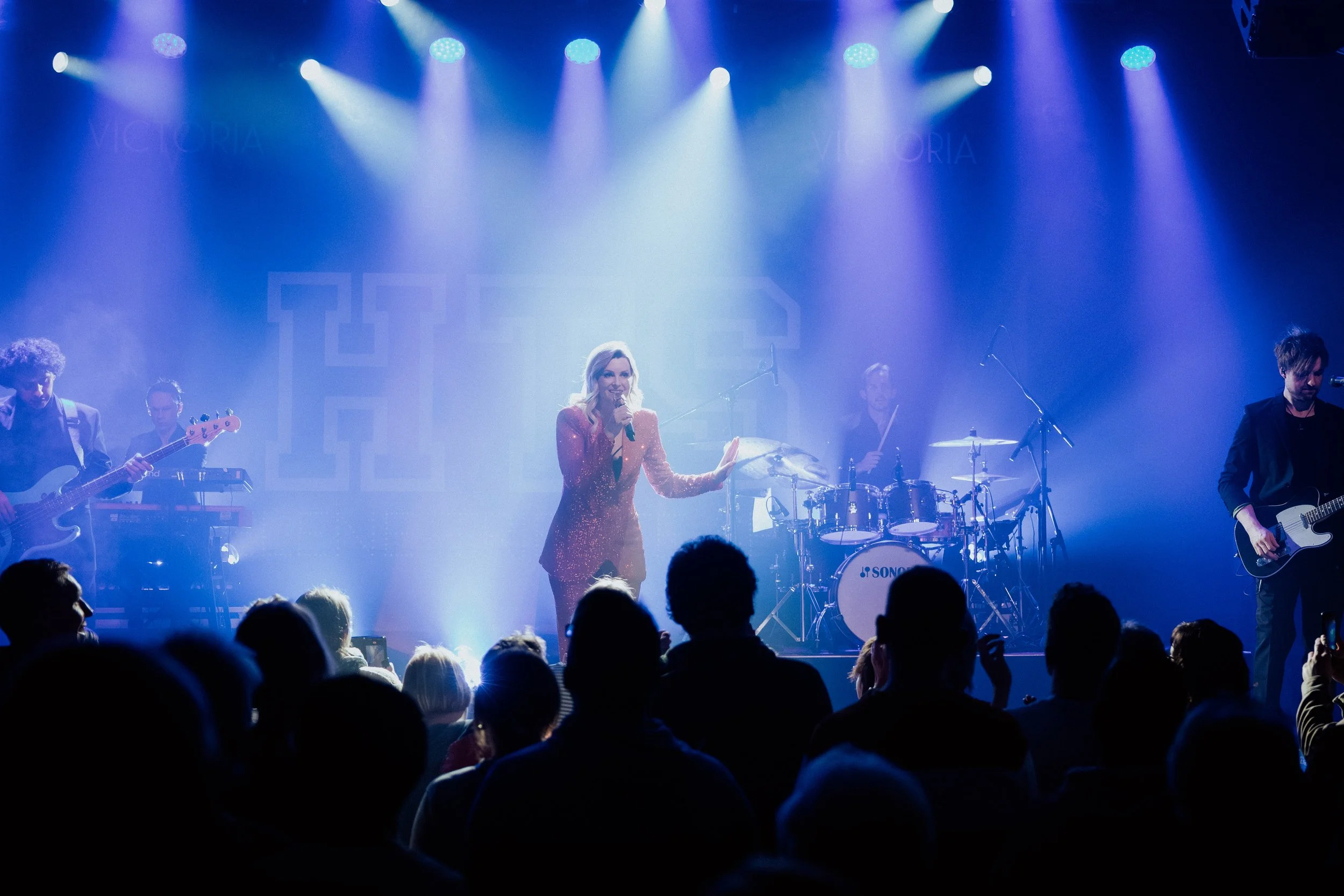 Eine Musikband tritt auf einer Bühne vor einem Publikum auf. Die Sängerin trägt ein rotes, glitzerndes Kleid und hält ein Mikrofon. Es gibt verschiedene Instrumente, darunter Gitarre, Schlagzeug und Keyboard, und farbige Bühnenbeleuchtung.