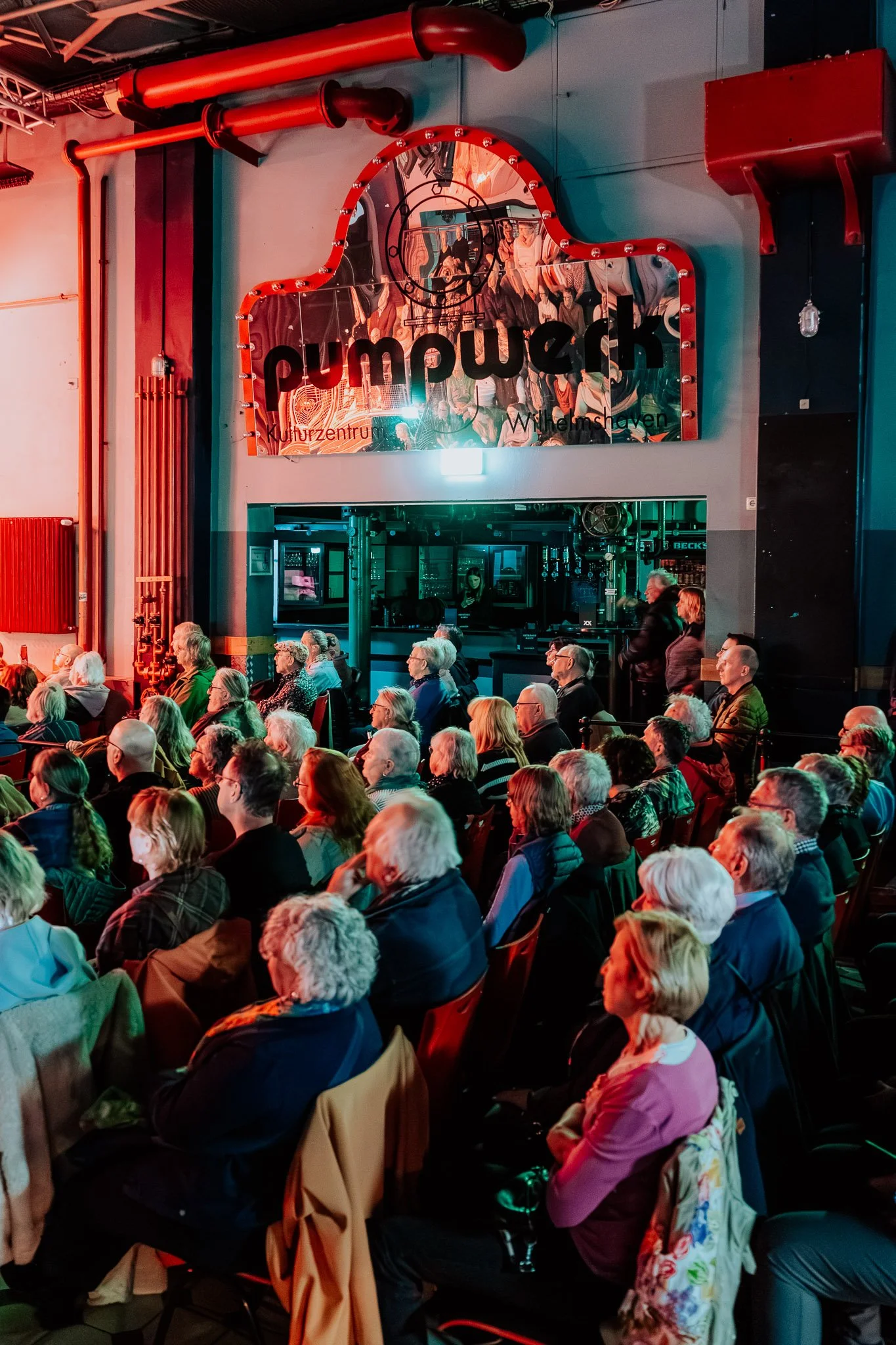 Ein Saal mit vielen älteren Menschen, die auf Stühlen sitzen, während eine Veranstaltung oder Aufführung stattfindet. Es gibt ein großes Wandbild mit dem Schriftzug 'pumuckl Kultzentrum Wilhelmshaven' im Hintergrund.