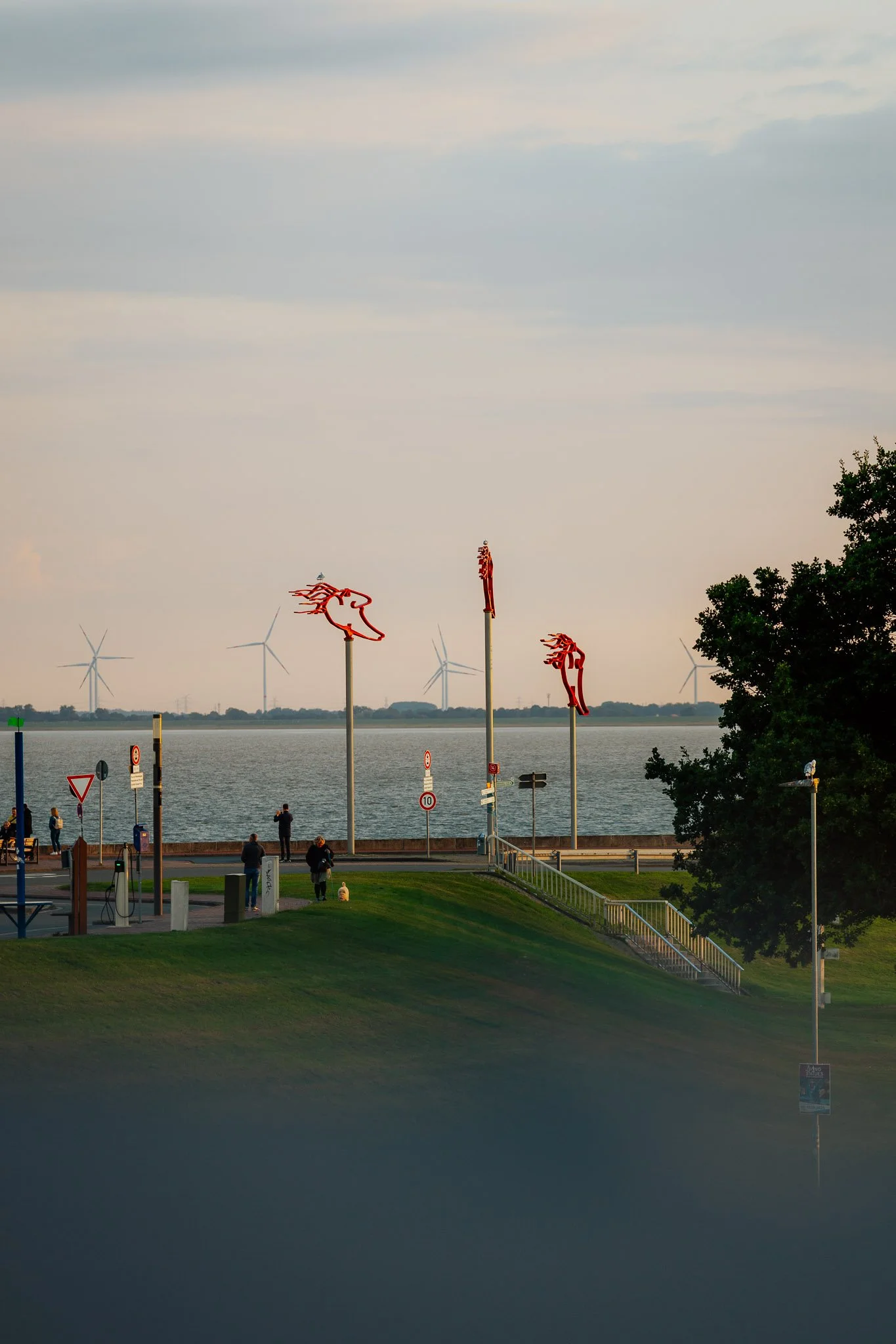 Menschen am Flussufer, Windräder im Hintergrund, Himmel mit Wolken, einige Steh- und Sitzgelegenheiten, Bäume und eine Wiese im Vordergrund, Abenddämmerung.