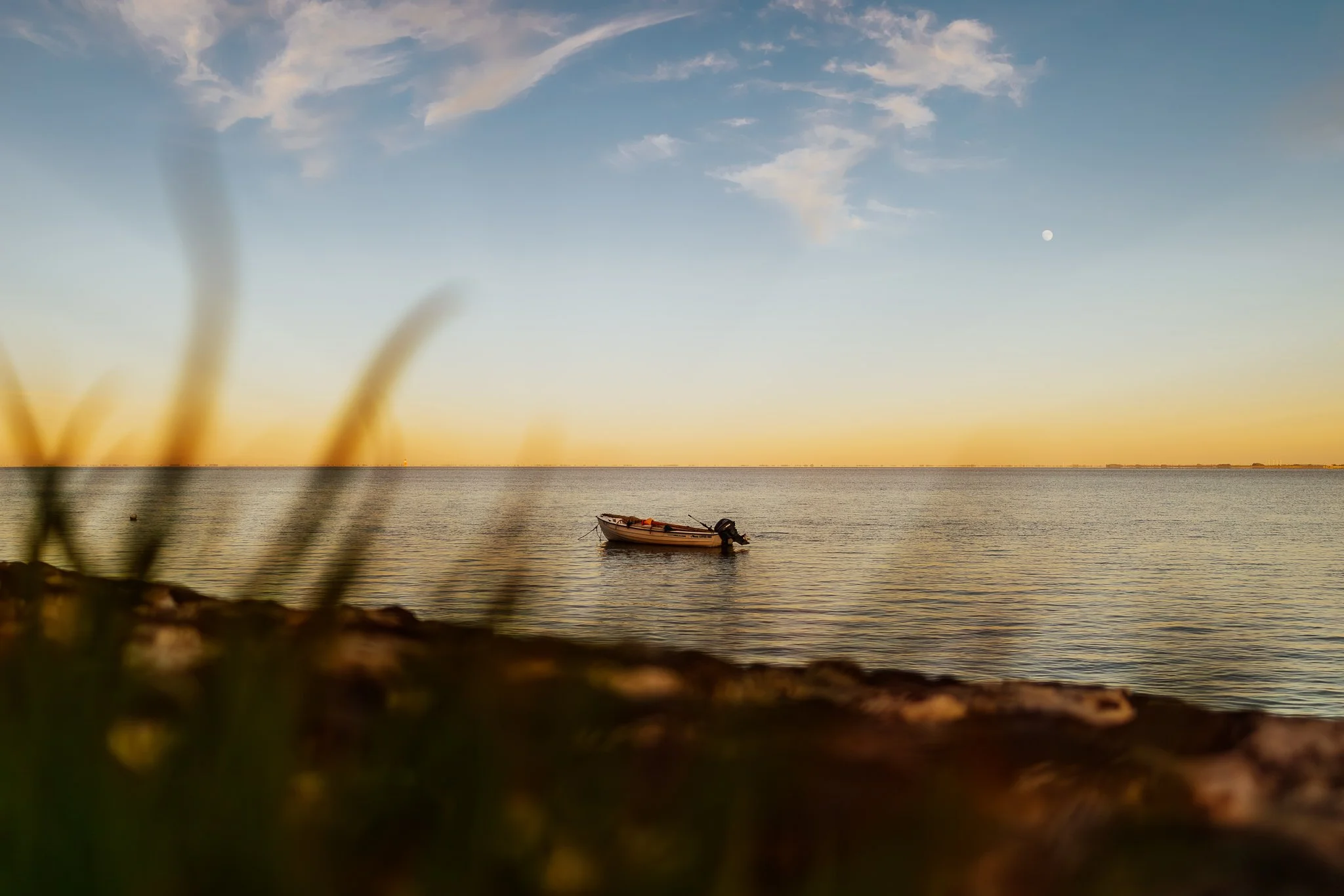 Ein Boot schwimmt auf ruhigem Wasser vor der Küste bei Sonnenuntergang, mit einem Mond am Himmel, im Vordergrund unscharfe Gräser.