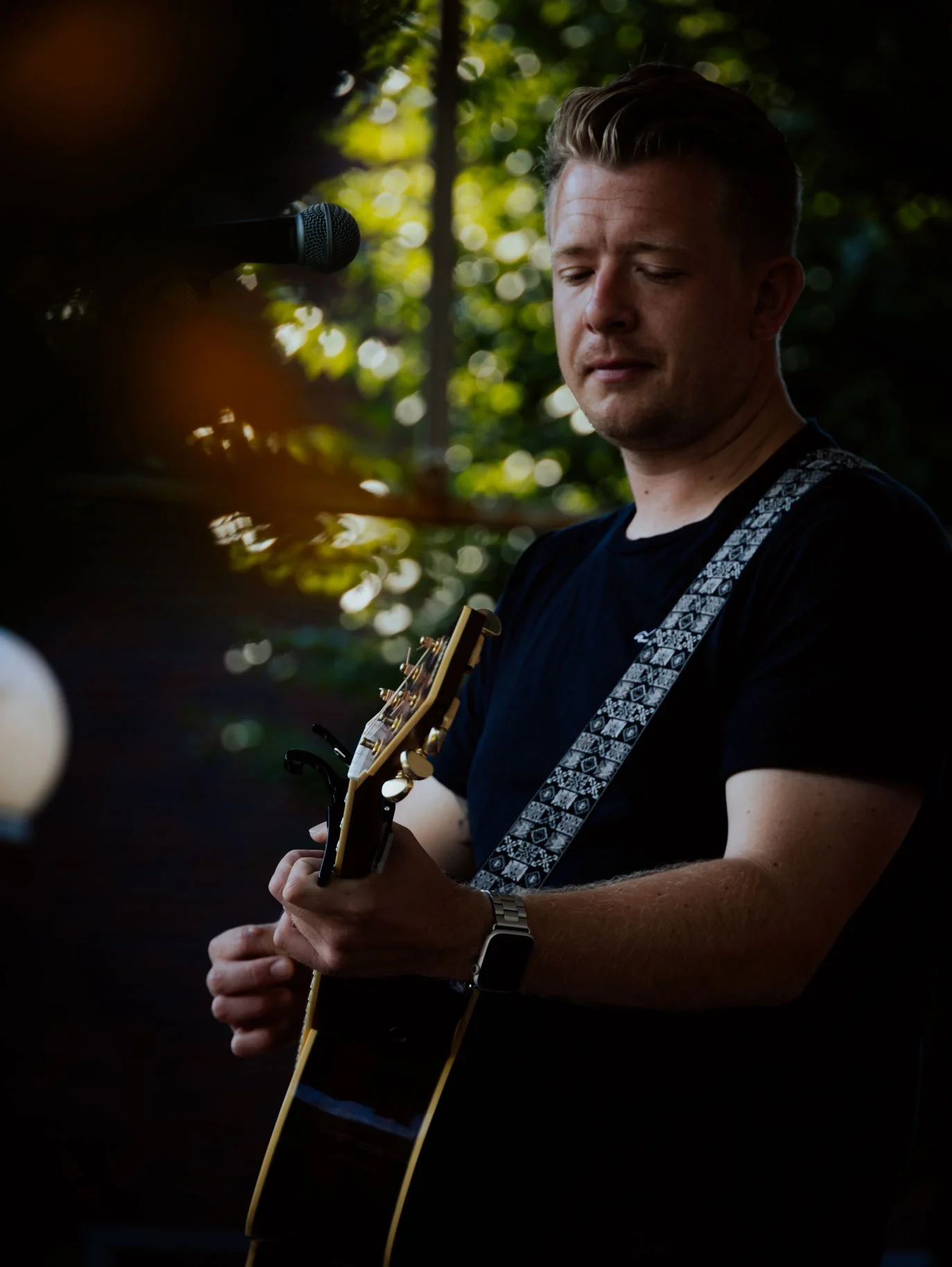 Ein Mann spielt Gitarre im Freien bei Sonnenlicht, während er vor einem Mikrofon steht.