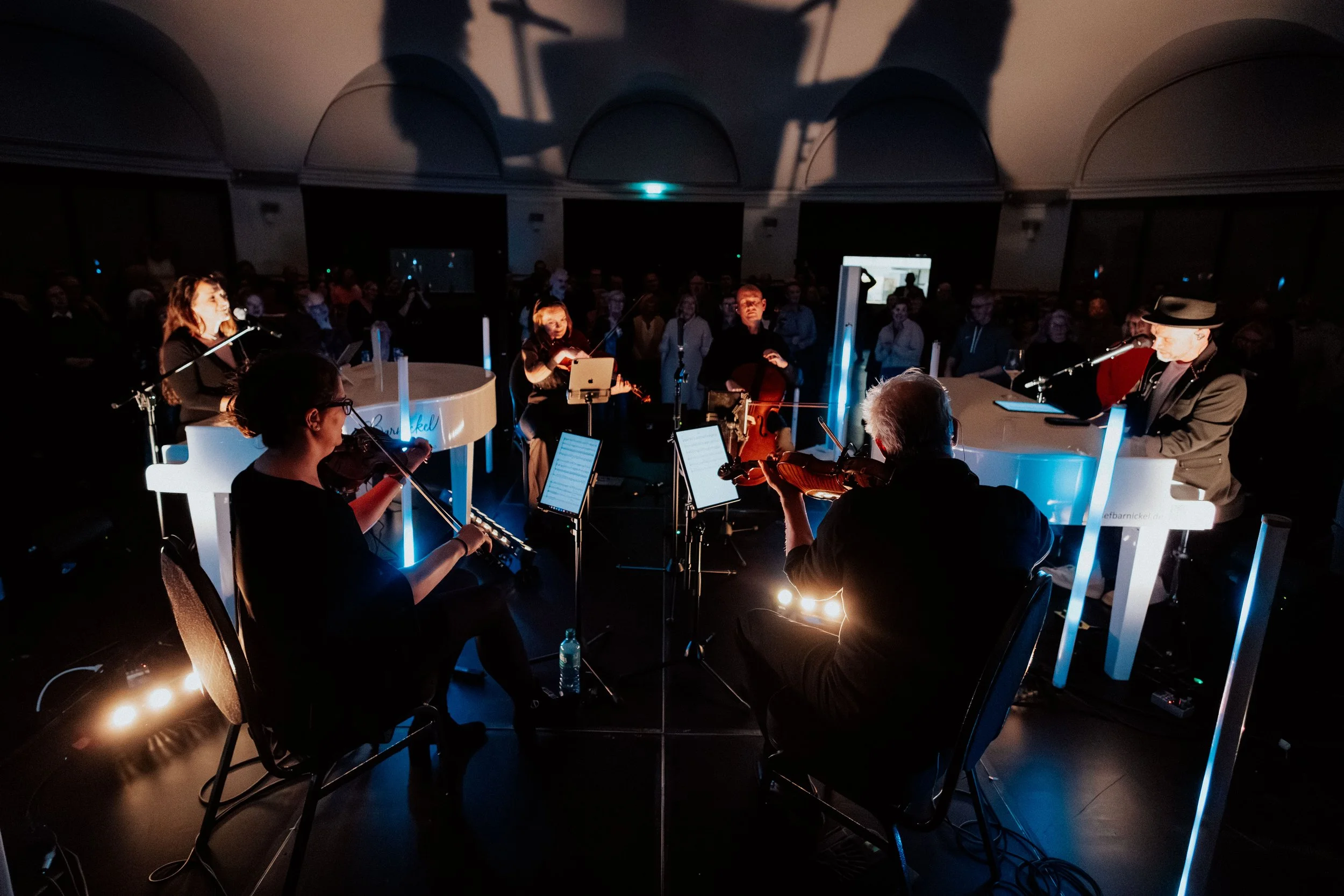 Musiker spielen in einem dunklen Saal, umgeben von einem Publikum, bei einem Konzert, beleuchtet von blauen Lichteffekten.