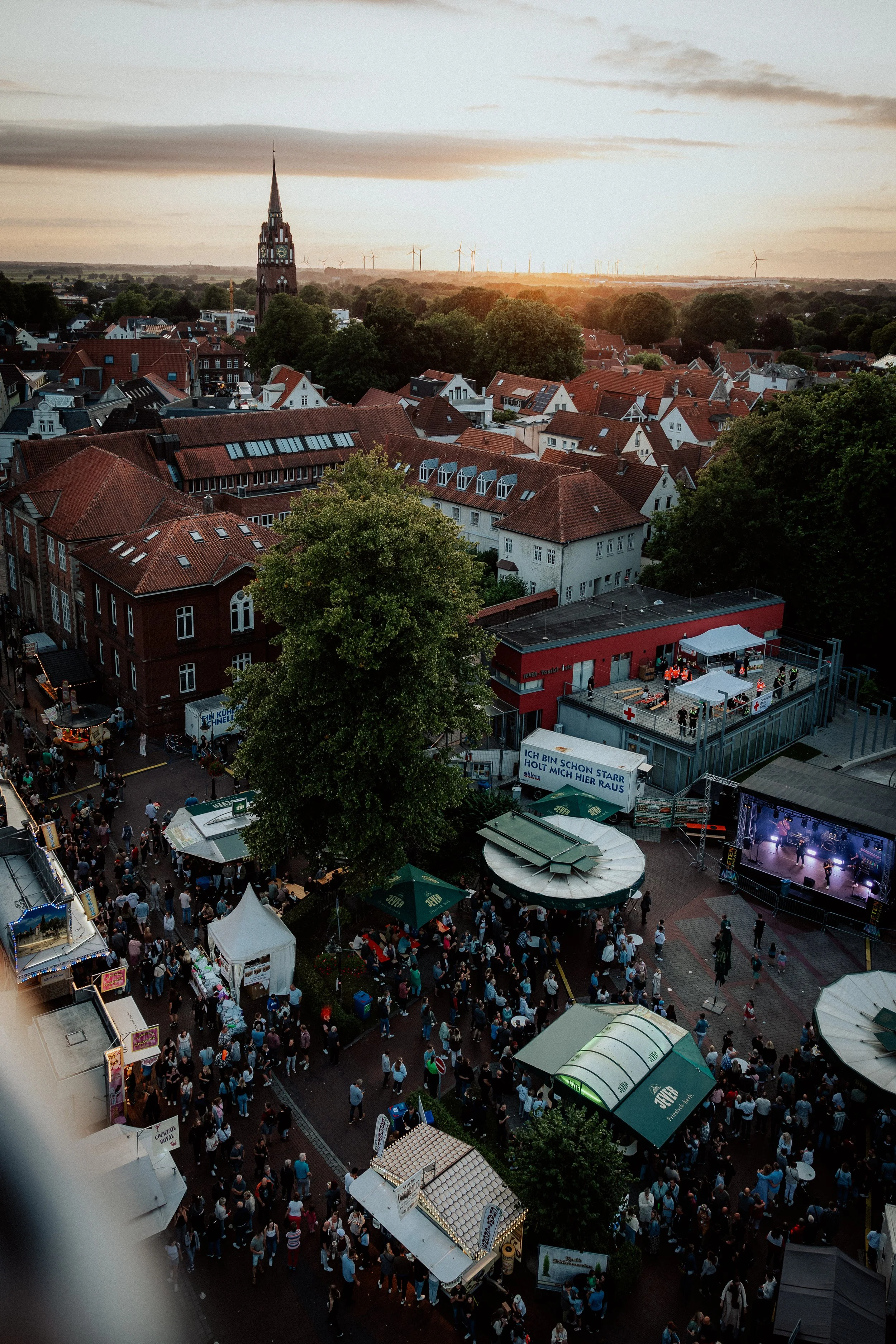 Blick auf ein Fest mit vielen Menschen, Ständen und einer Bühne, bei Sonnenuntergang in einer Stadt mit Kirchturm und Windrädern im Hintergrund.