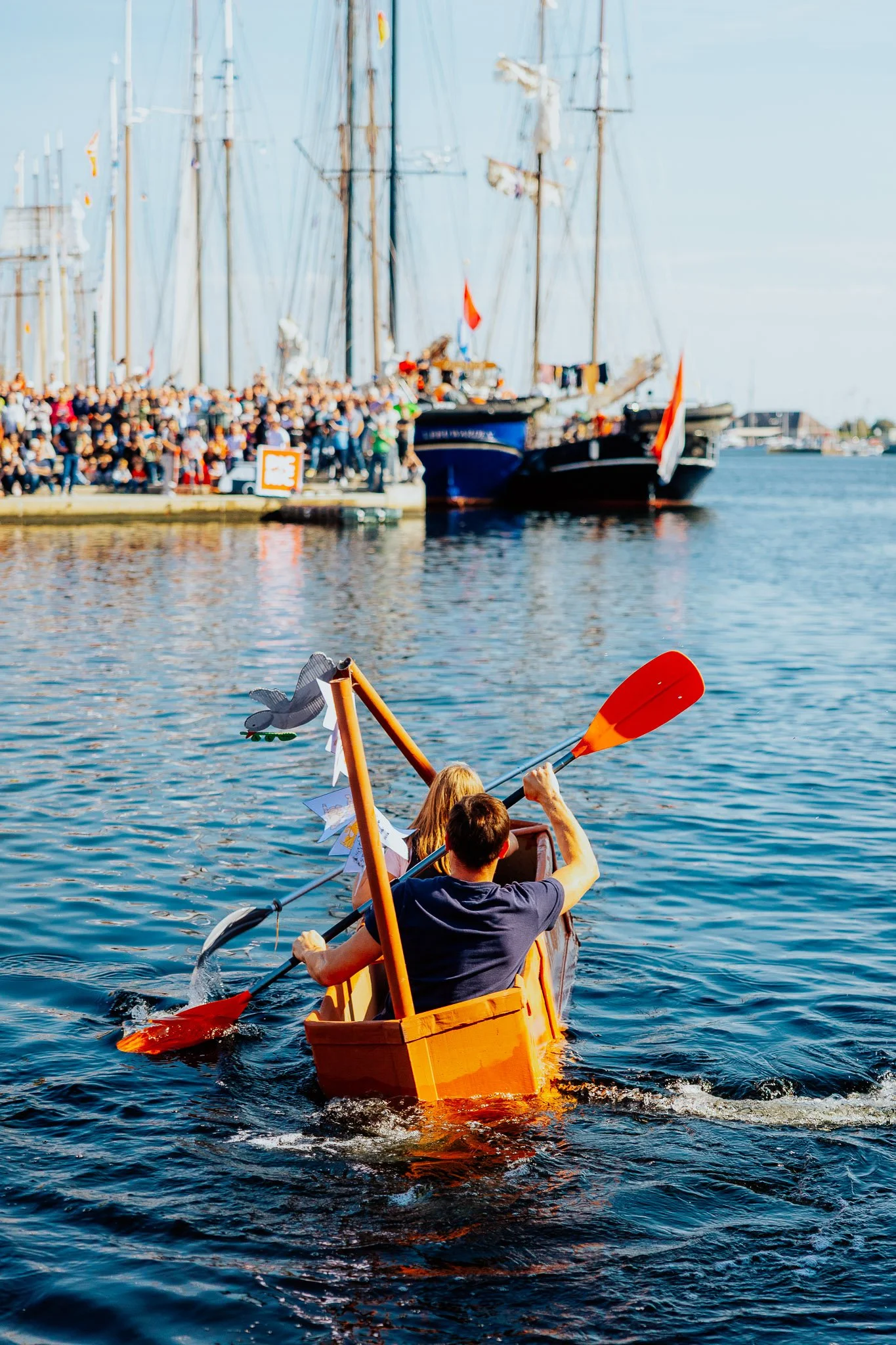 Zwei Personen paddeln in einem orangefarbenen Boot auf dem Wasser, im Hintergrund sind große Segelboote und eine Menschenmenge am Ufer sichtbar.