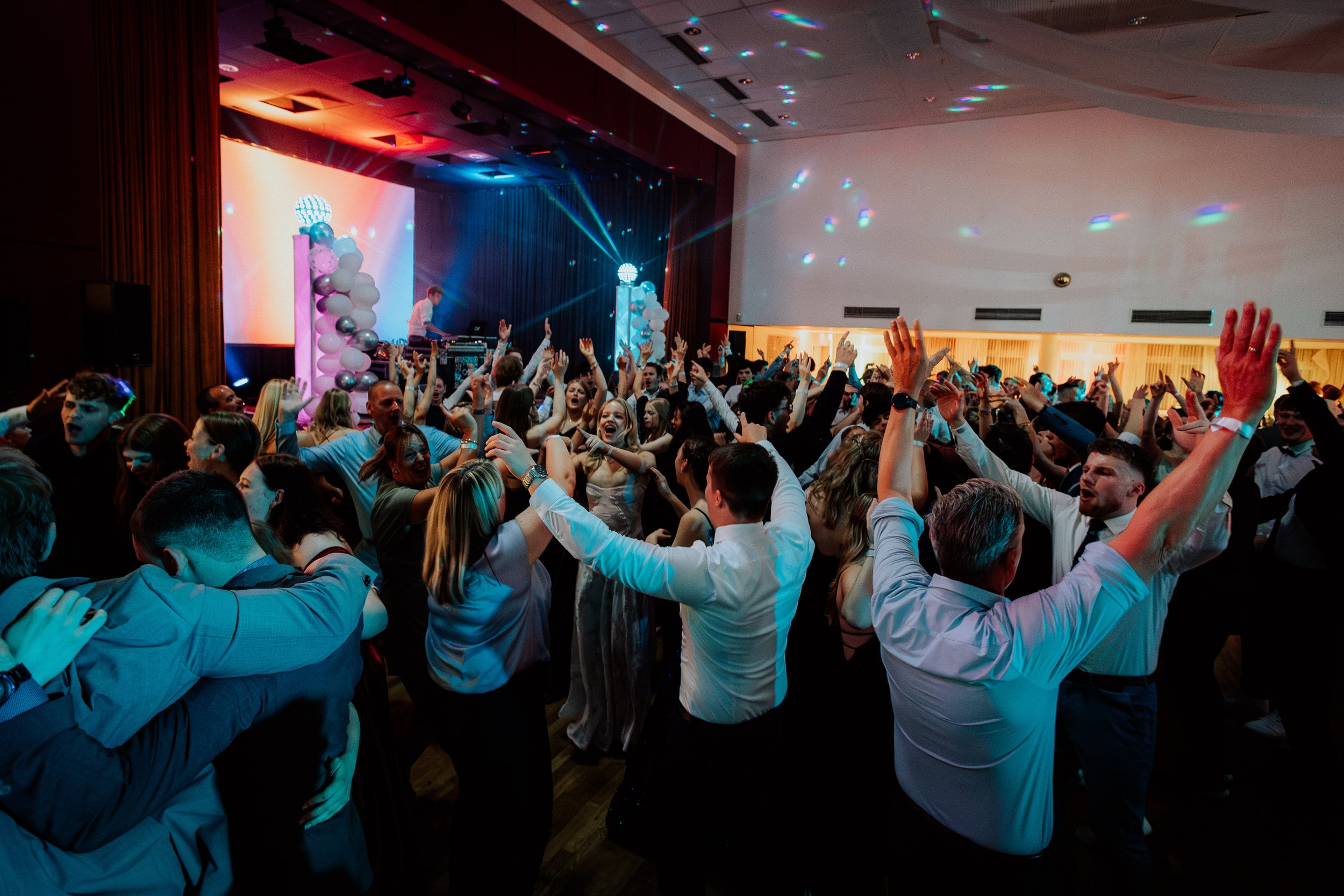 Menschen tanzen und feiern auf einer Party im festlich dekorierten Raum mit bunten Lichteffekten, auf einer Bühne steht ein DJ.