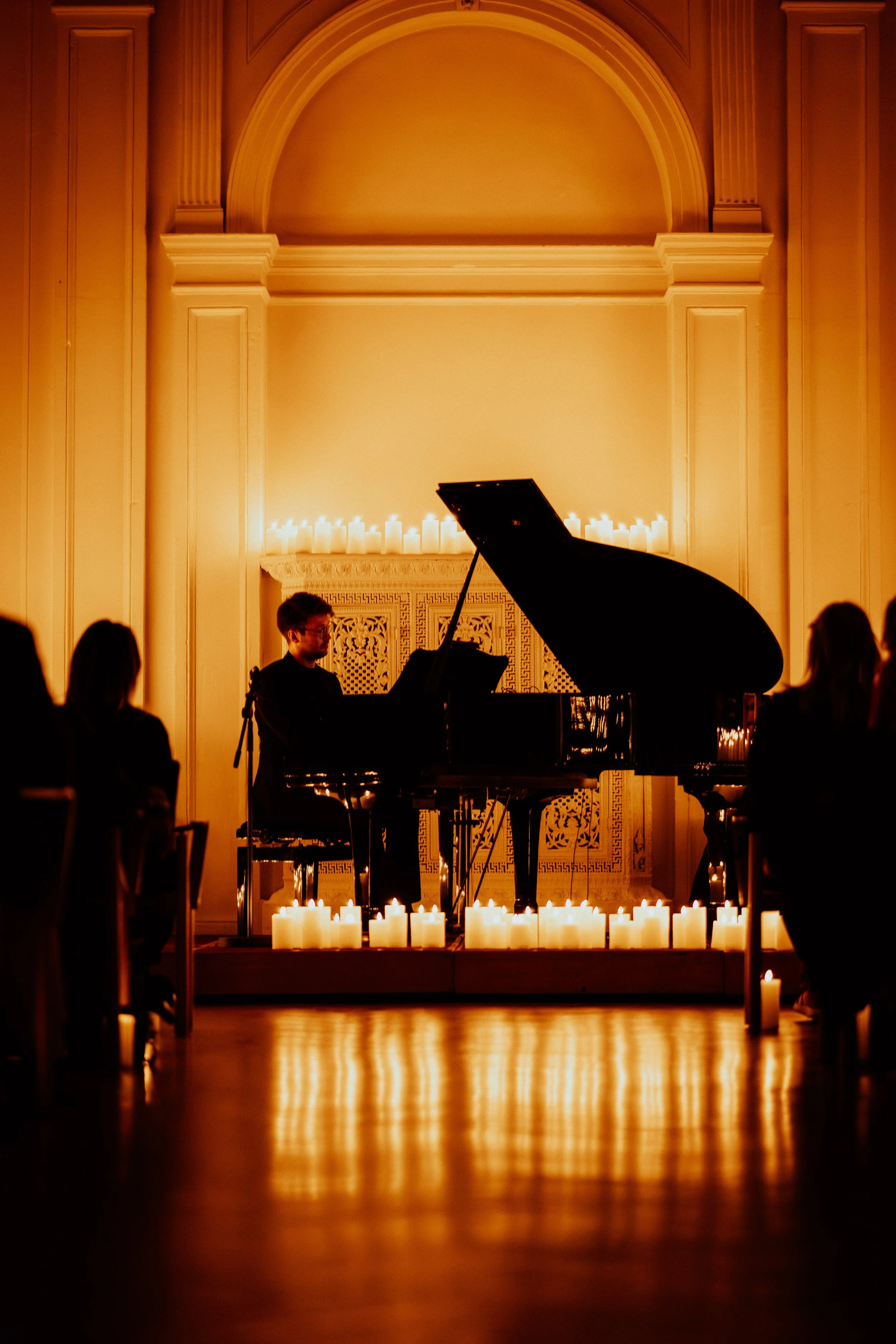 Ein Musiker spielt Klavier in einem Raum mit Kerzen, die den Hintergrund und den Boden beleuchten, während das Publikum im Dunkeln sitzt.