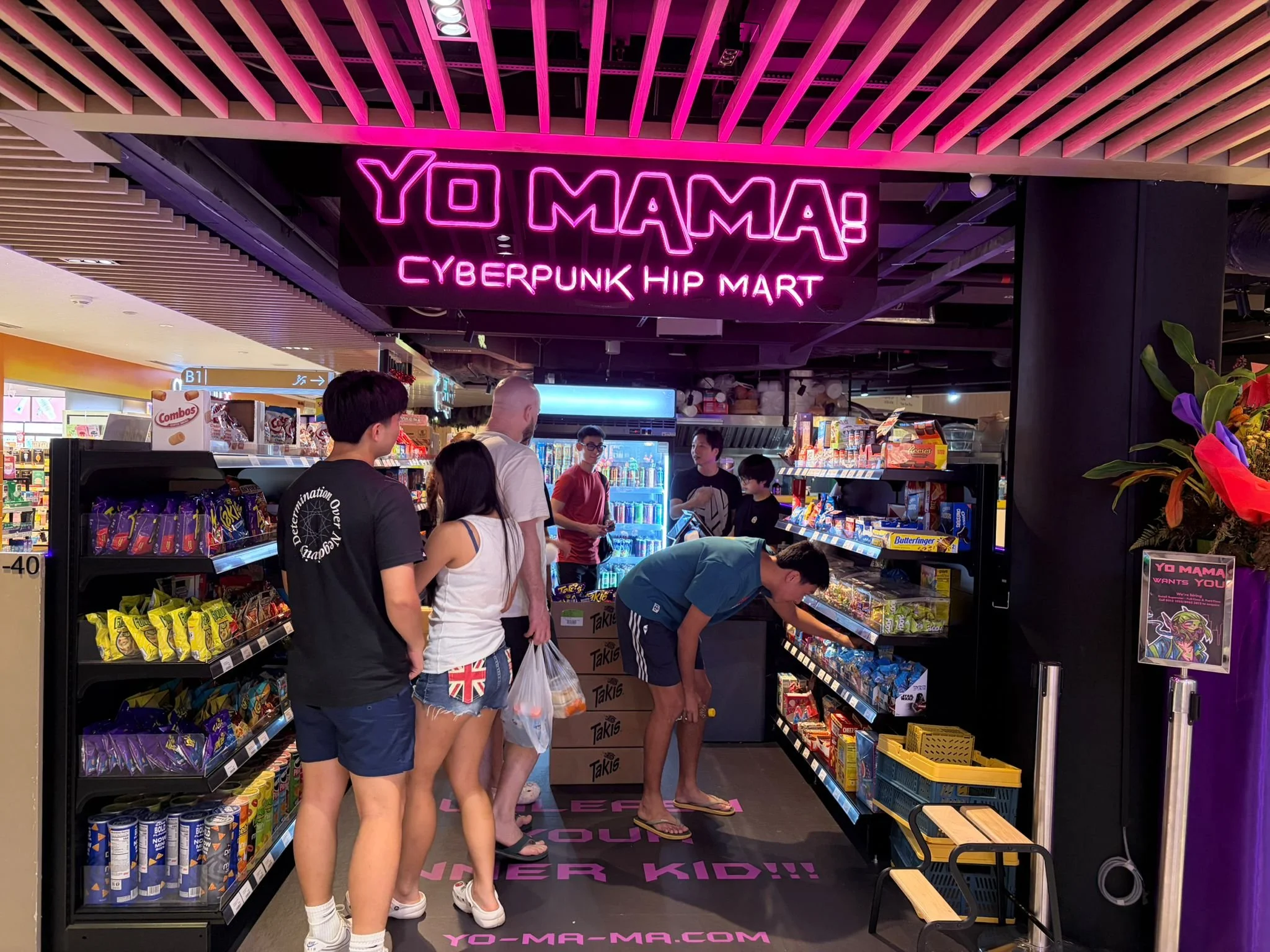 People shopping at a store with a neon sign that reads ''YO MAMA! CYBERPUNK HIP MART'' in pink lights, surrounded by shelves of snacks and snacks in colorful packaging.