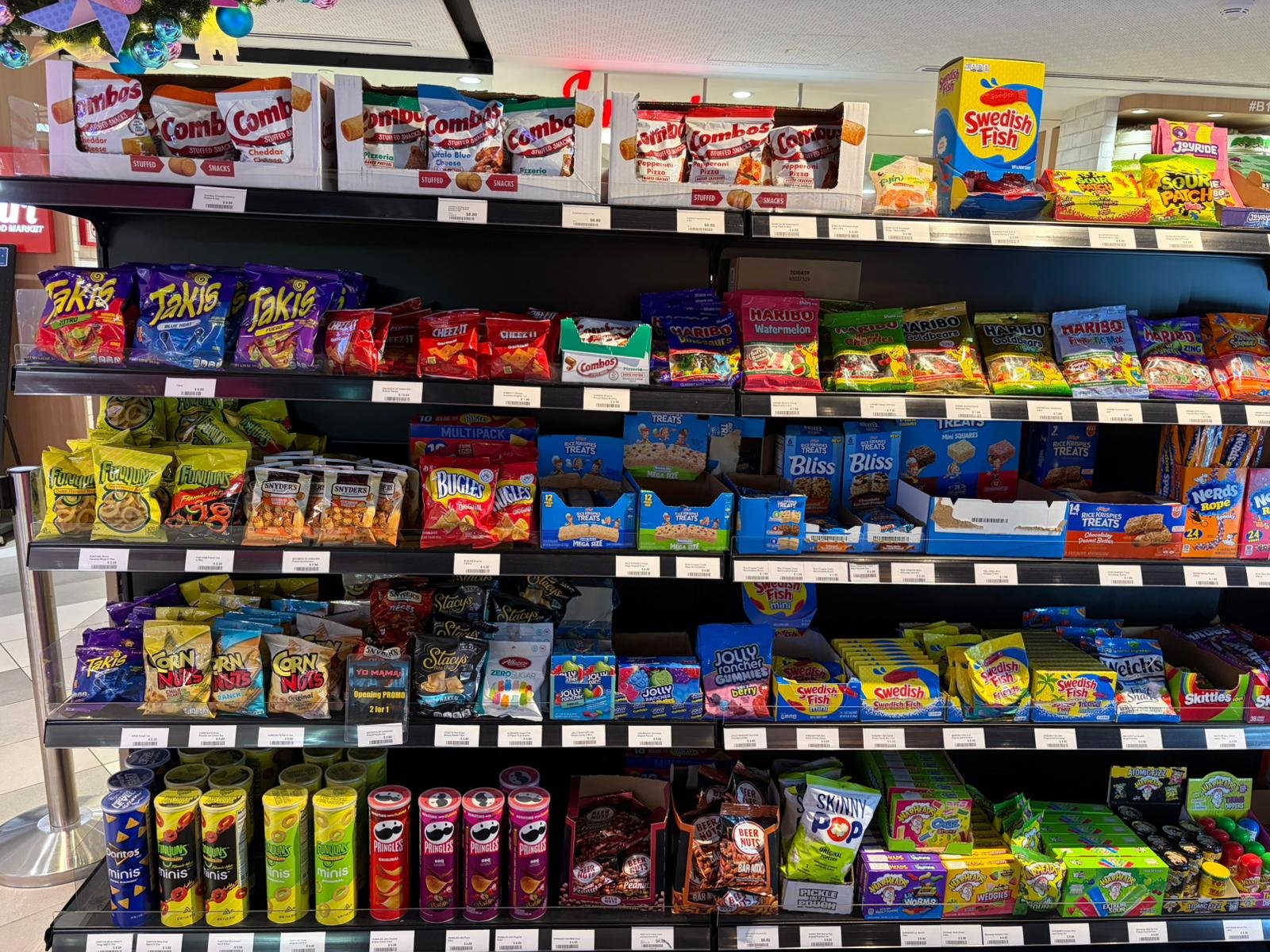 Shelf display in a store with various snack foods including bags of Combos, Takis, Swiss Fish candies, Sour Patch, Nerds Rope, Pringles, and other candies and chips.