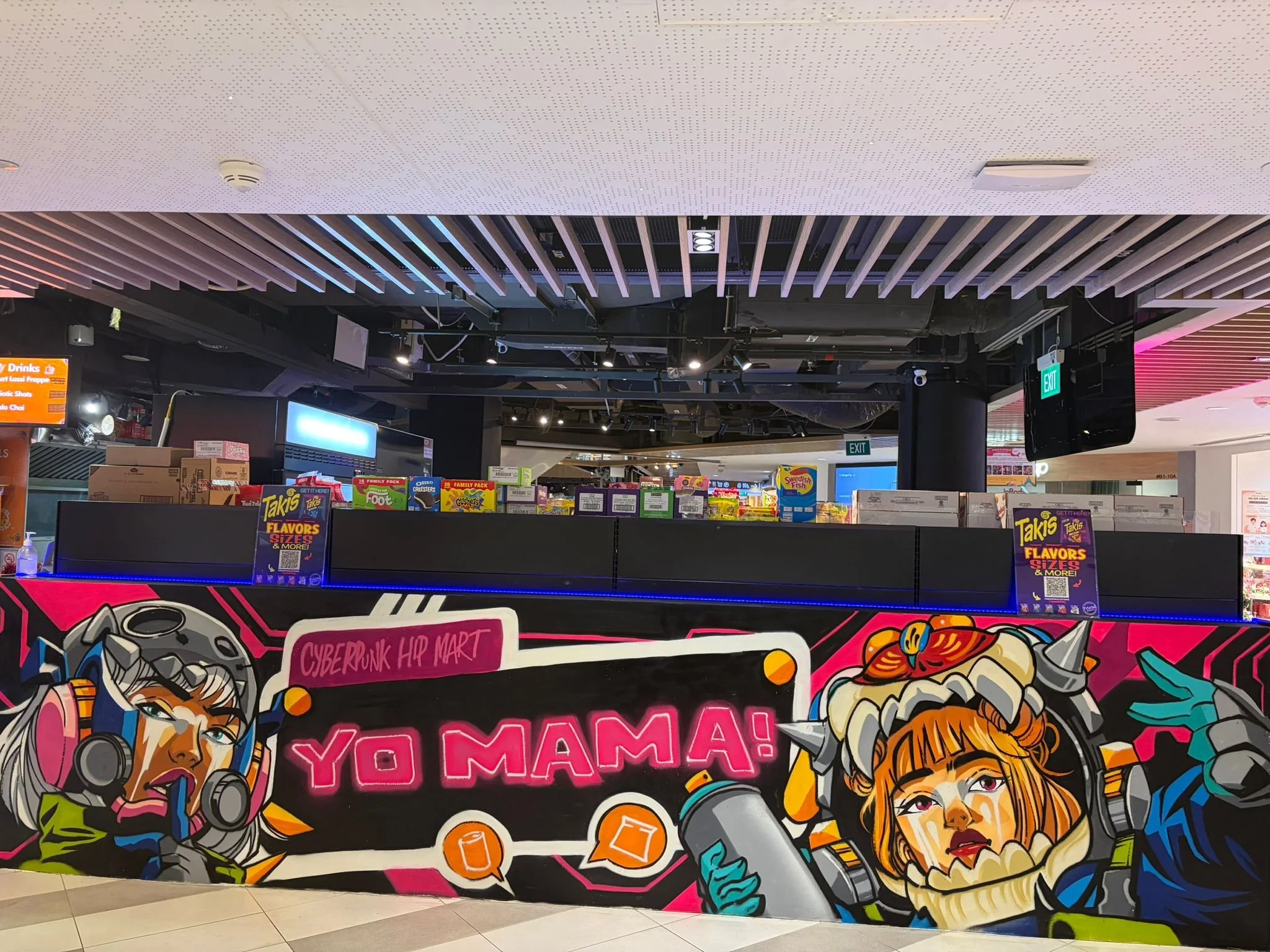 Colorful graffiti art on a display board with illustrations of women in futuristic outfits, and neon text reading 'YumYum! YO MAMA!' in a retail store.