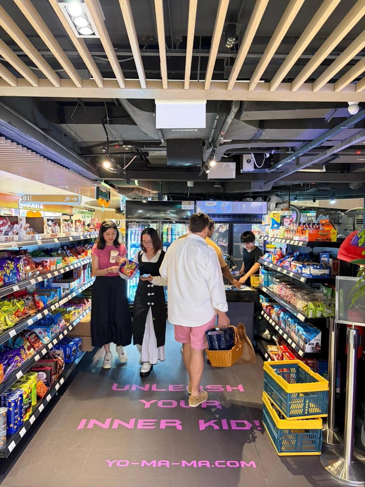 Inside a convenience store with shoppers browsing shelves filled with snacks and candies, with a sign on the floor reading 'Unleash Your Inner Kid!'