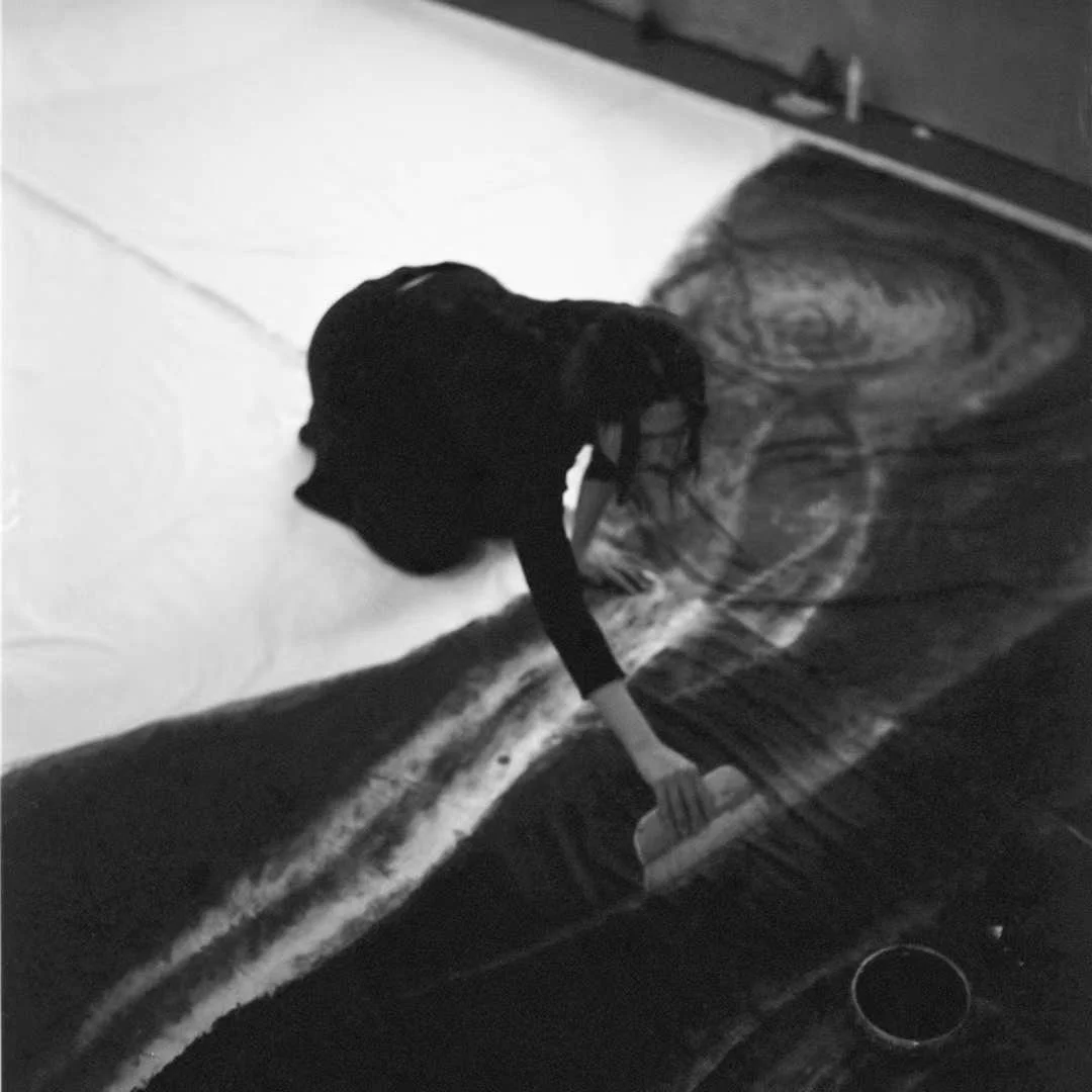 A person in black clothing is cleaning or arranging a bed with a patterned comforter and white sheets in a bedroom.
