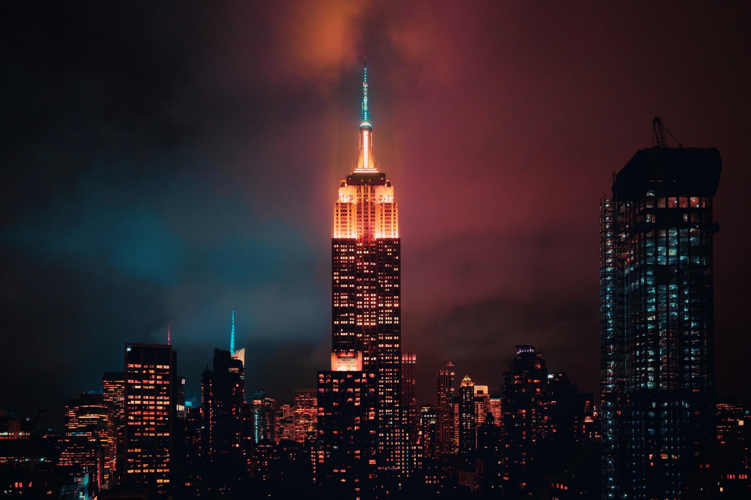 Nighttime view of the Empire State Building illuminated with red and orange lights, surrounded by other lit skyscrapers in New York City against a cloudy sky.