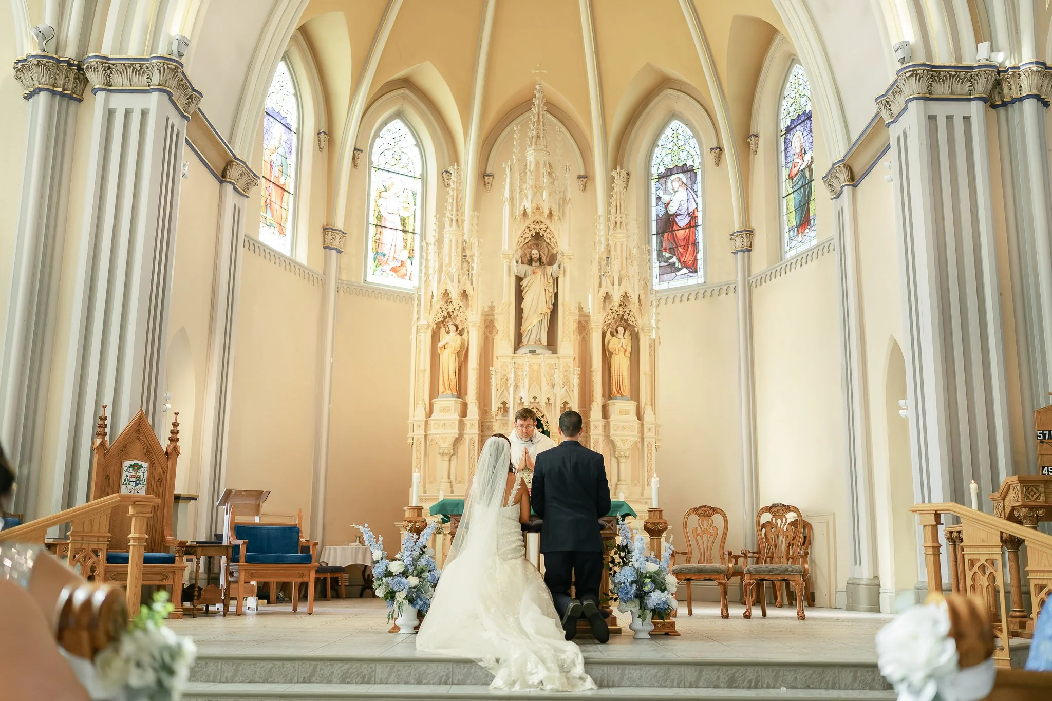 A bride and groom kneeling at the altar of a church during their wedding ceremony. The bride is wearing a white wedding dress with a veil, and the groom is in a dark suit. The church has tall stained-glass windows and ornate religious statues and decorations at the altar.