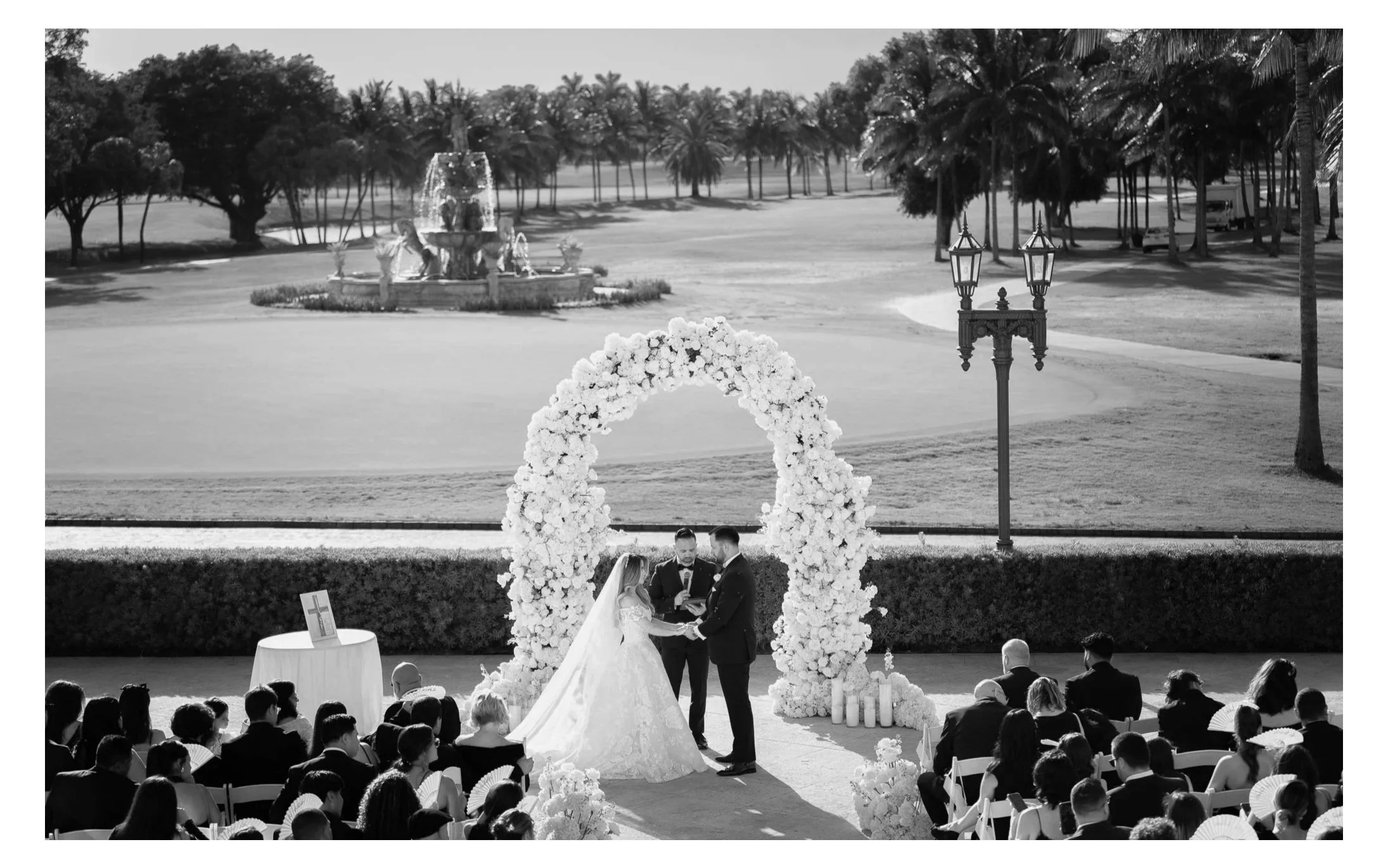 A black and white photo of a wedding ceremony outdoors on a sunny day, with a bride and groom holding hands under a floral arch, surrounded by seated guests, a table with an icon, and a scenic background of trees, a fountain, and a golf course.
