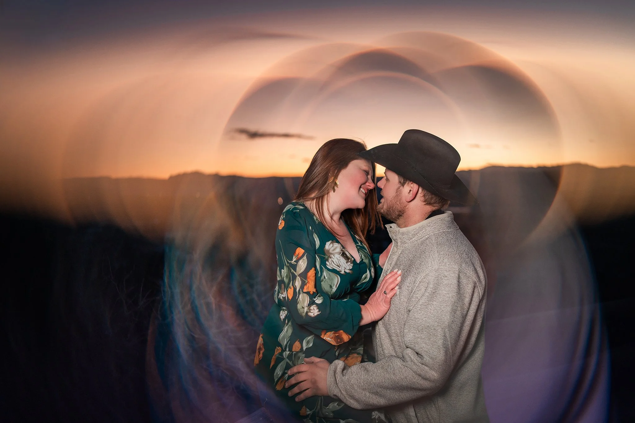 A couple, a woman and a man, are close together and smiling at each other during sunset, with a large heart-shaped lens flare framing them.