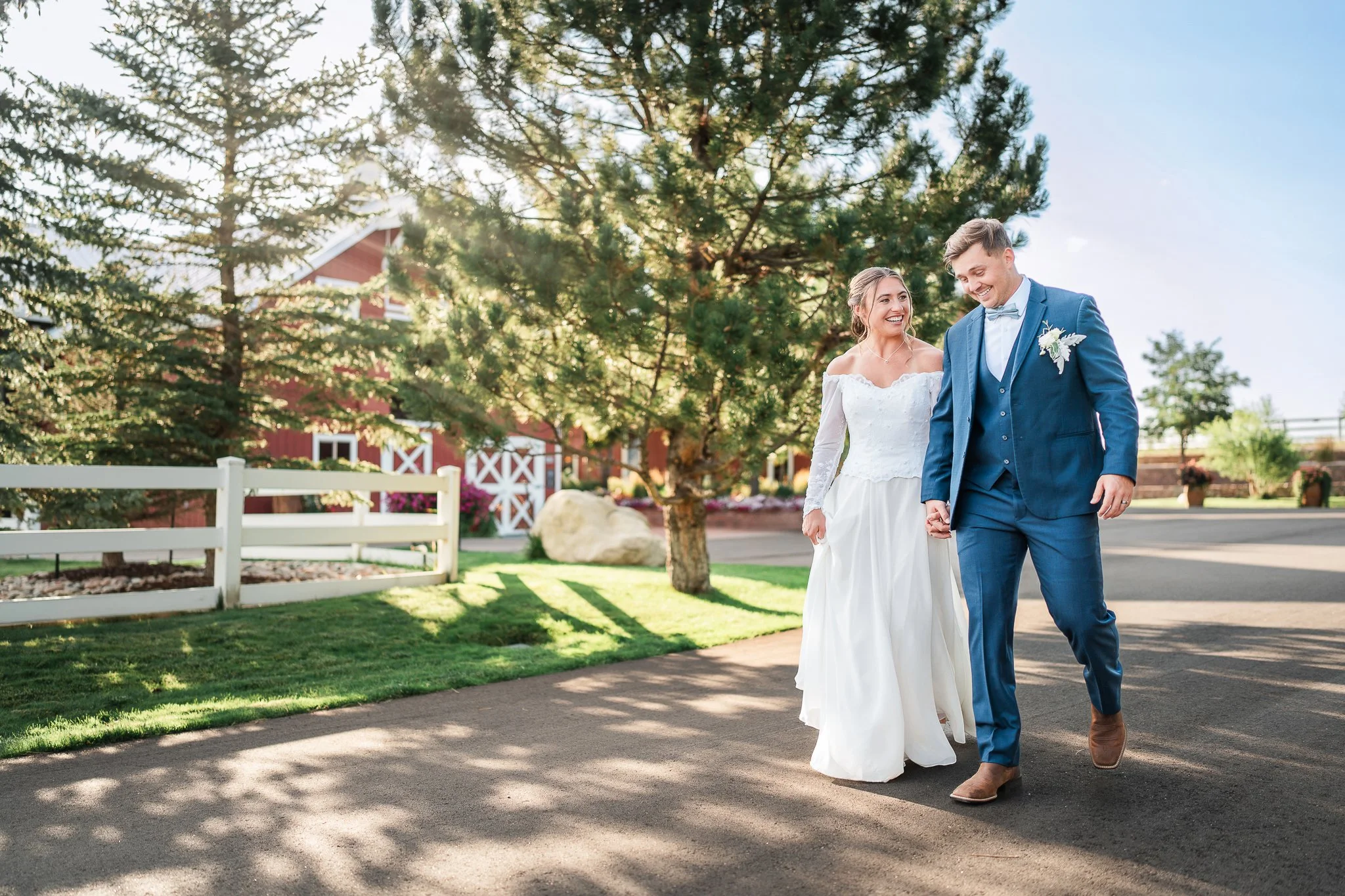 A bride and groom holding hands and walking on a paved path outdoors during daytime, with trees and a red barn-like building in the background.