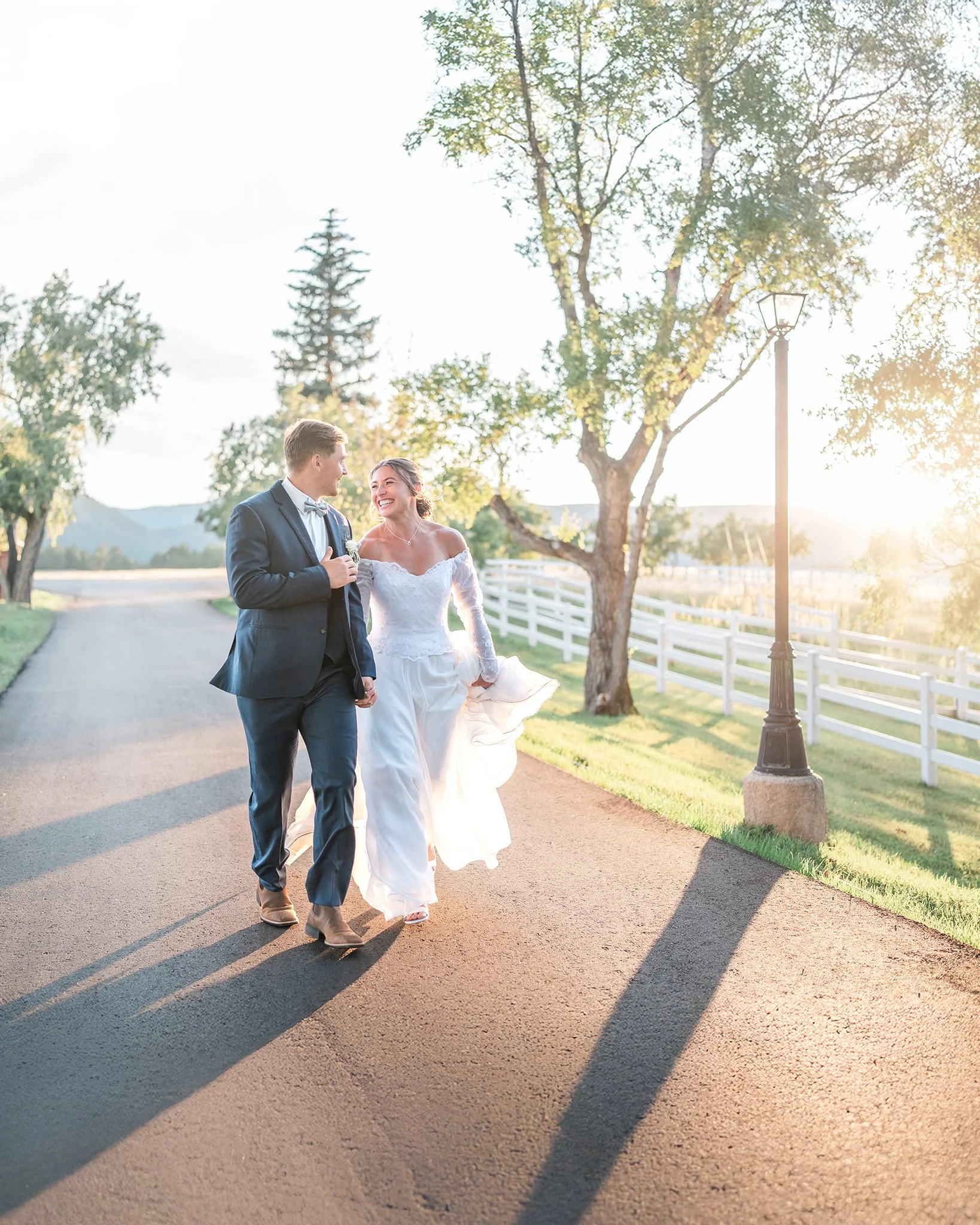 A newlywed couple walking on a paved road in the countryside during sunset, smiling at each other, with trees, a white fence, and a lamp post in the background.