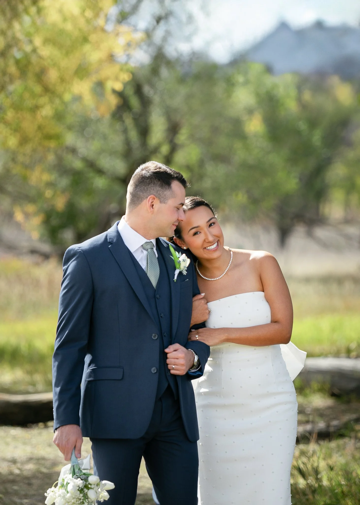 A couple in wedding attire outdoors, with the woman smiling and leaning her head on the man's shoulder, surrounded by trees and natural scenery.