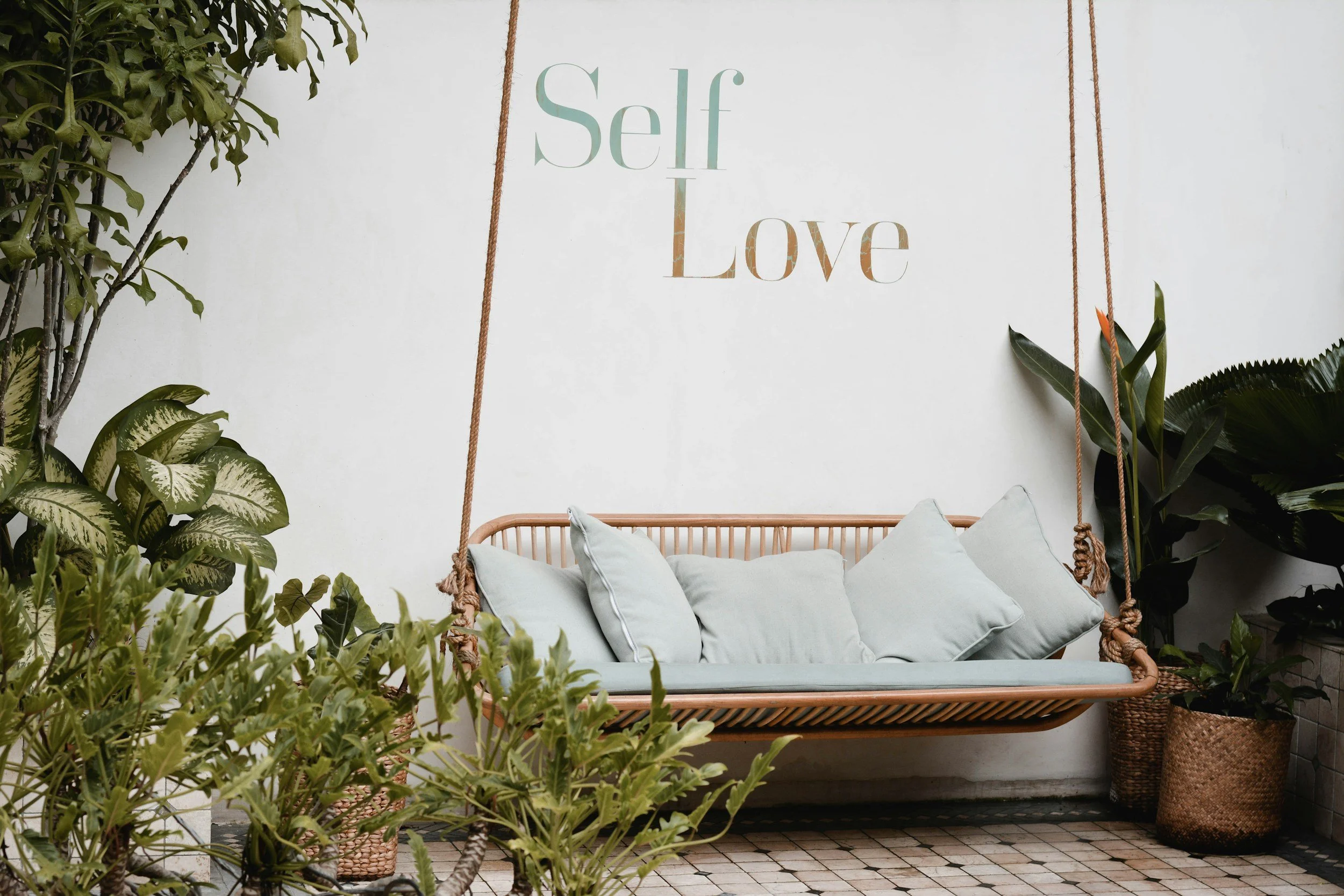 A hanging wooden swing with white cushions, surrounded by green plants, and a white wall in the background with the words 'Self Love' written on it.