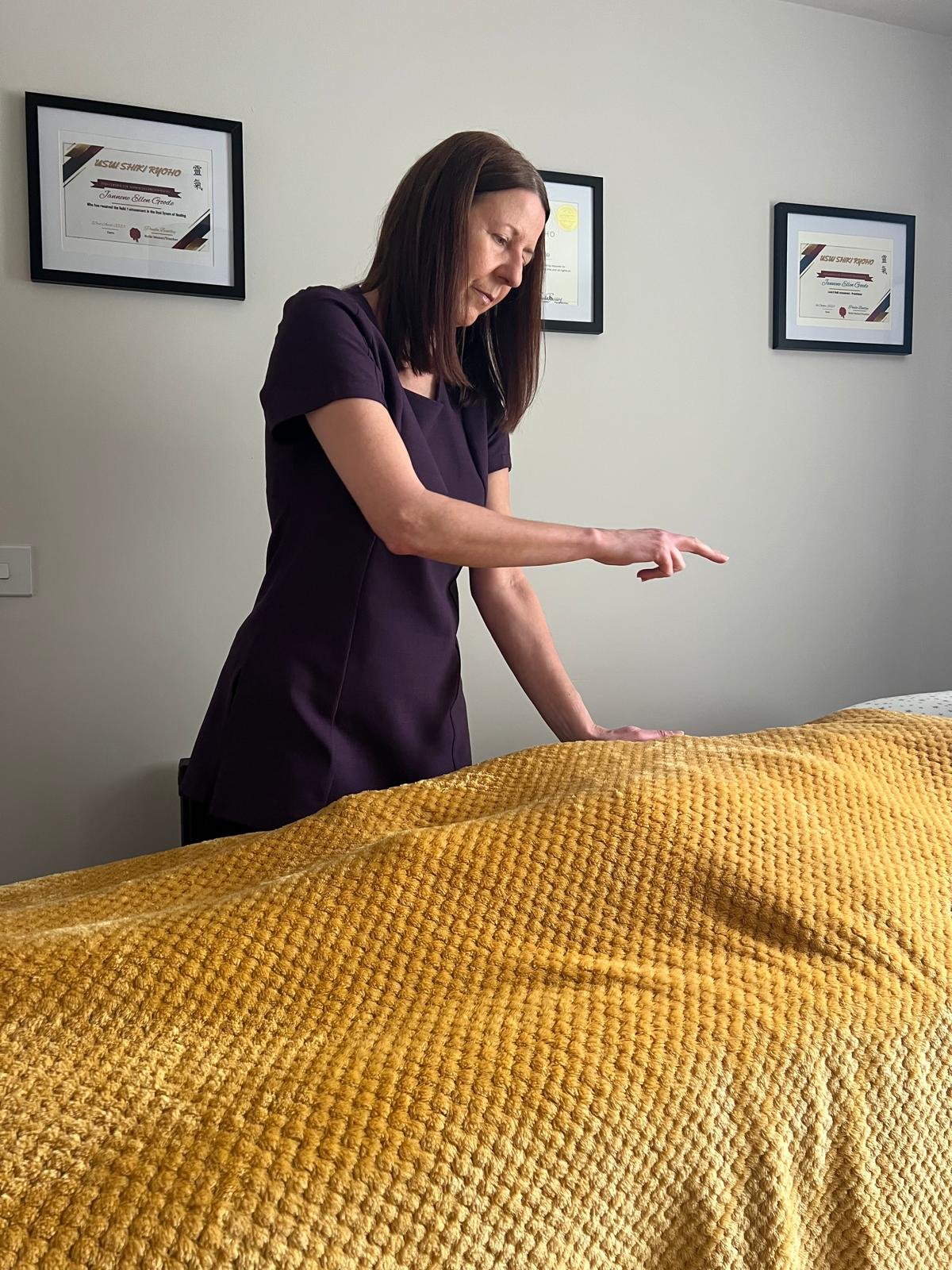 A woman in a dark purple uniform performing a massage on a client covered with a yellow textured blanket in a room with framed certificates on the wall.