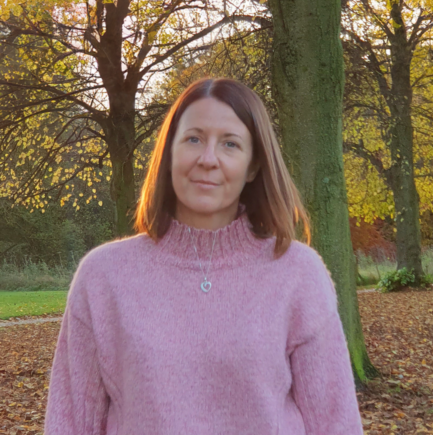 A woman with shoulder-length brown hair wearing a pink sweater and a heart-shaped pendant necklace standing outdoors near trees with autumn foliage.