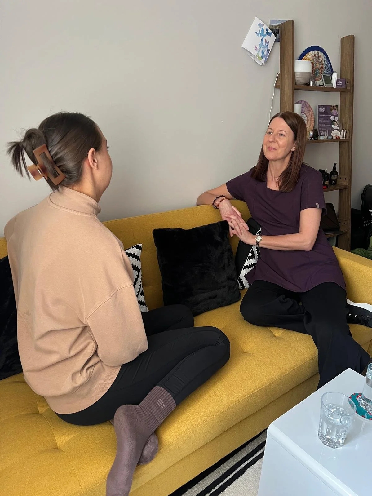 A woman in a purple dress sitting on a yellow couch, conversing with a woman in a beige sweatshirt and black pants, in a living room with a bookshelf in the background.