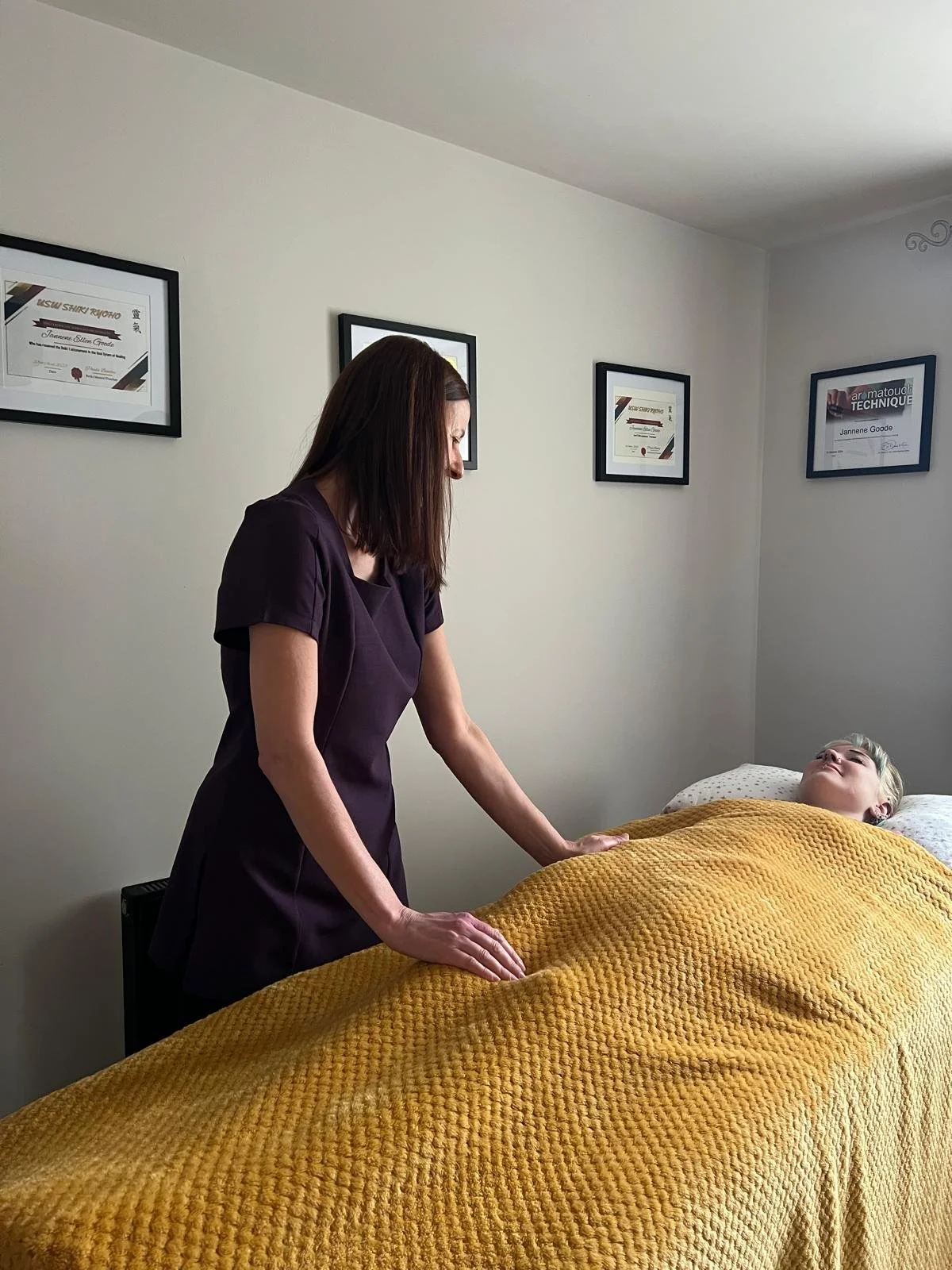 A woman receives a massage from a massage therapist in a treatment room with diplomas on the wall.