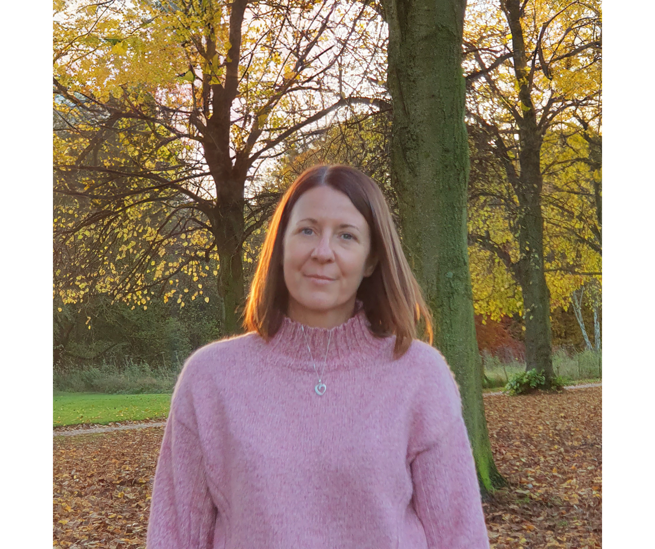 A woman with red hair taking a selfie outdoors in a park during autumn. She is wearing a pink fuzzy sweater and a necklace with a heart-shaped pendant. The background has trees with yellow leaves and a sunny sky.