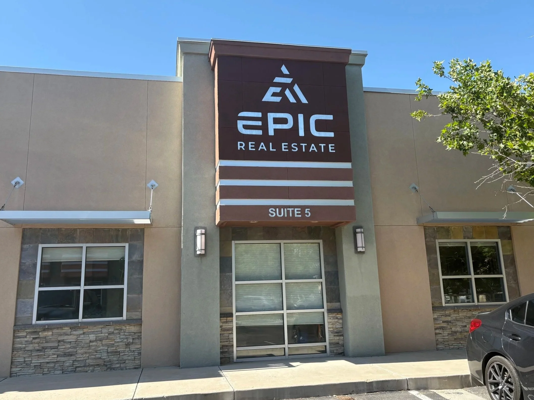 Exterior view of a commercial building with a sign that reads 'Epic Real Estate' and 'Suite 5'. The building has large windows, a mix of stone and stucco facade, and a parking lot with a gray car nearby. A green leafy tree is visible on the right, and the sky is clear and blue.