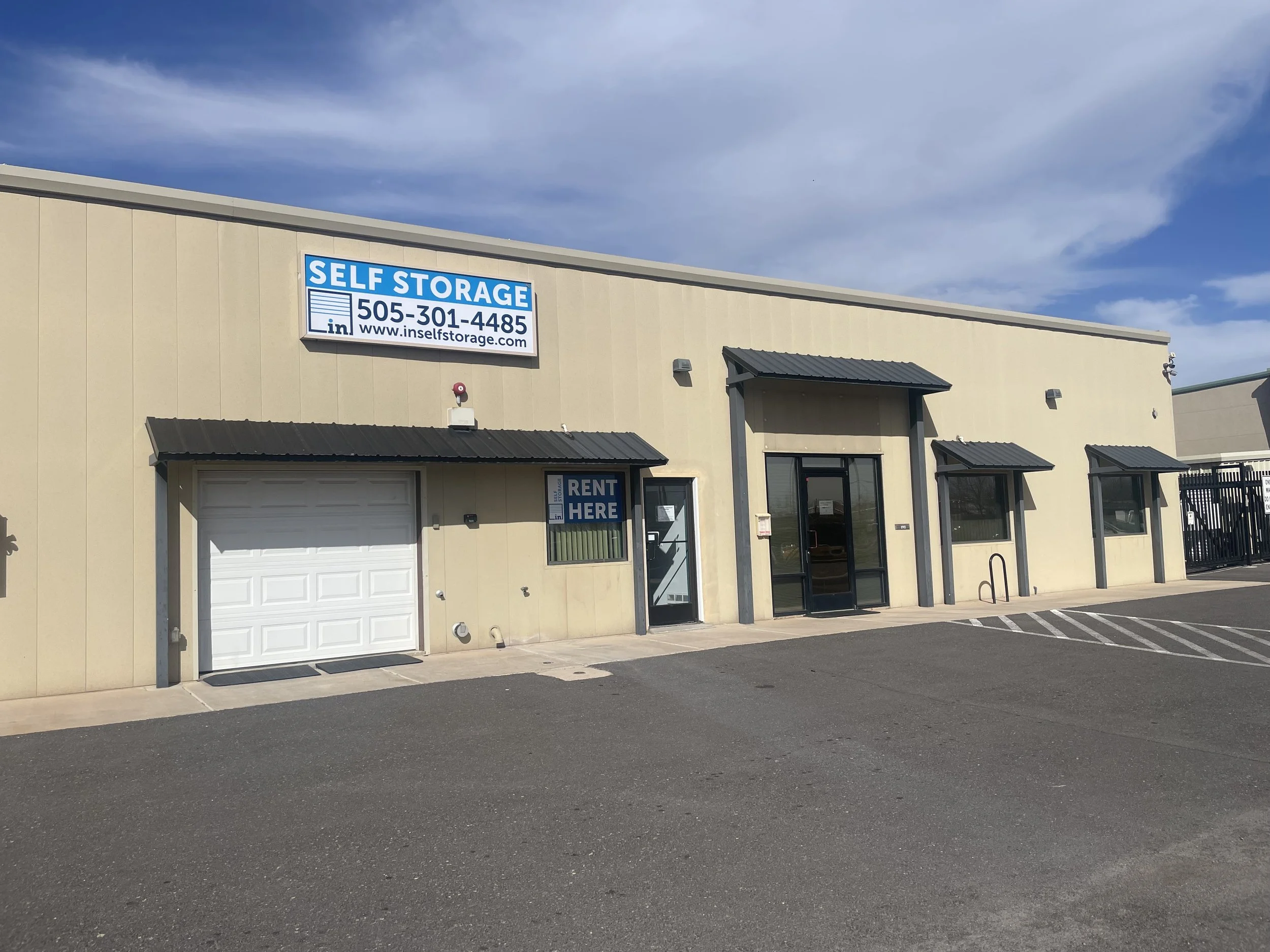 An exterior view of a beige commercial building with signs indicating it is a self-storage facility. There is a large sign with the text 'SELF STORAGE' and a phone number, along with a website address. The building has a garage door and a glass door with a small window, along with black awnings over the entrances. The pavement is marked with parking spaces, and there are a few security cameras and wall-mounted lights on the building. The sky is partly cloudy.