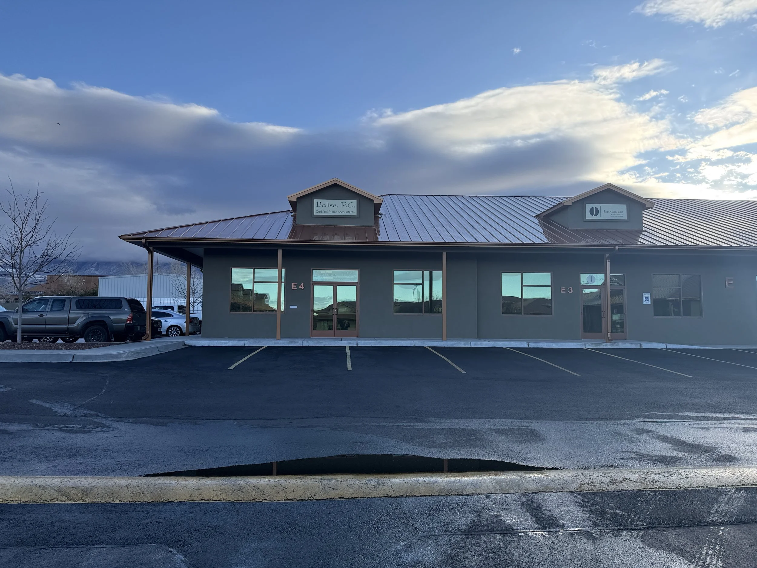 Front view of a commercial building with two signs on the roof, parking lot with cars, leafless trees, and a cloudy sky with some blue patches.