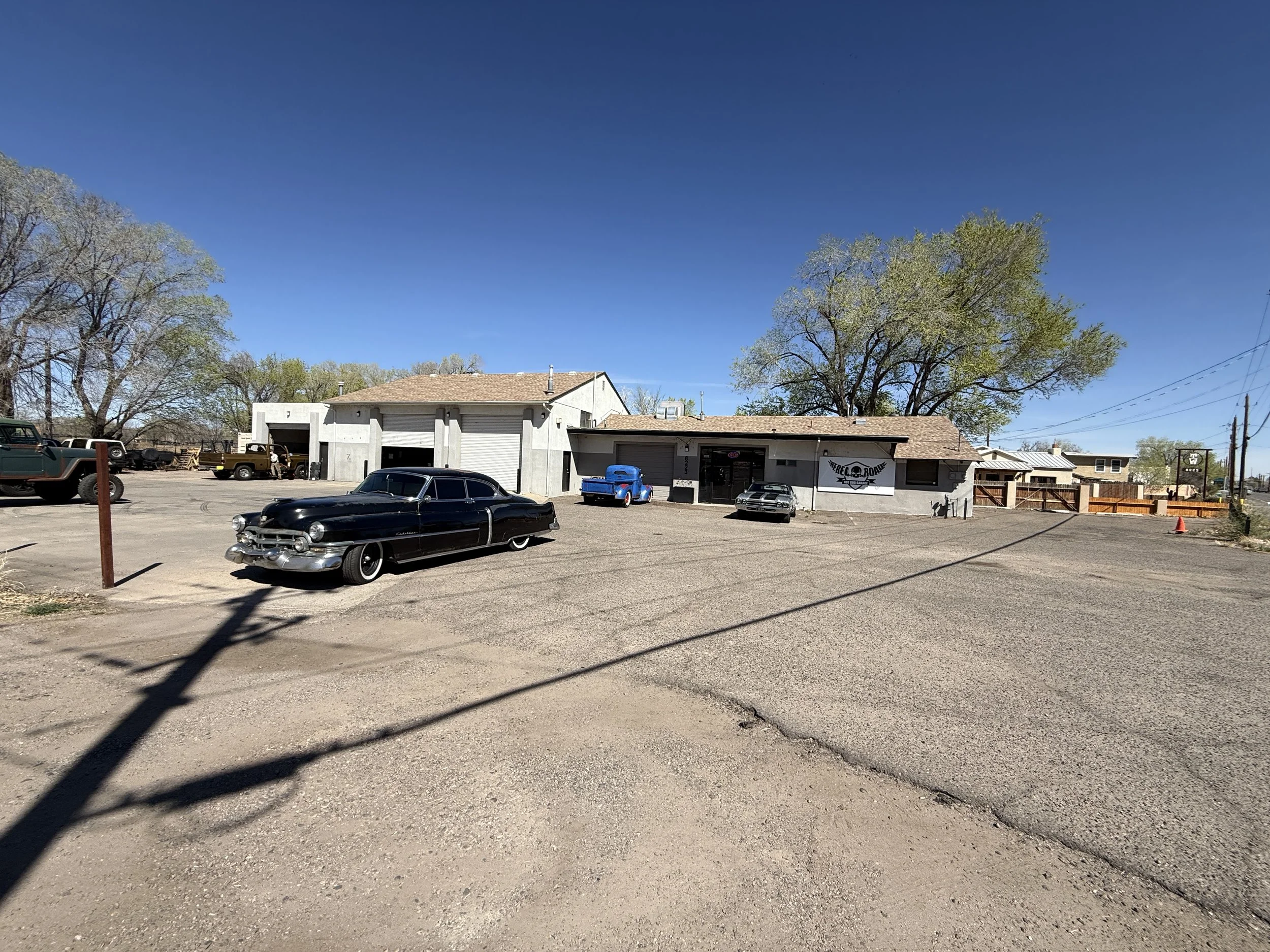 A parking lot with a graffiti shop in a white building, several vintage cars including a black Cadillac, a blue truck, and a black car parked outside on a sunny day with a clear blue sky and trees in the background.