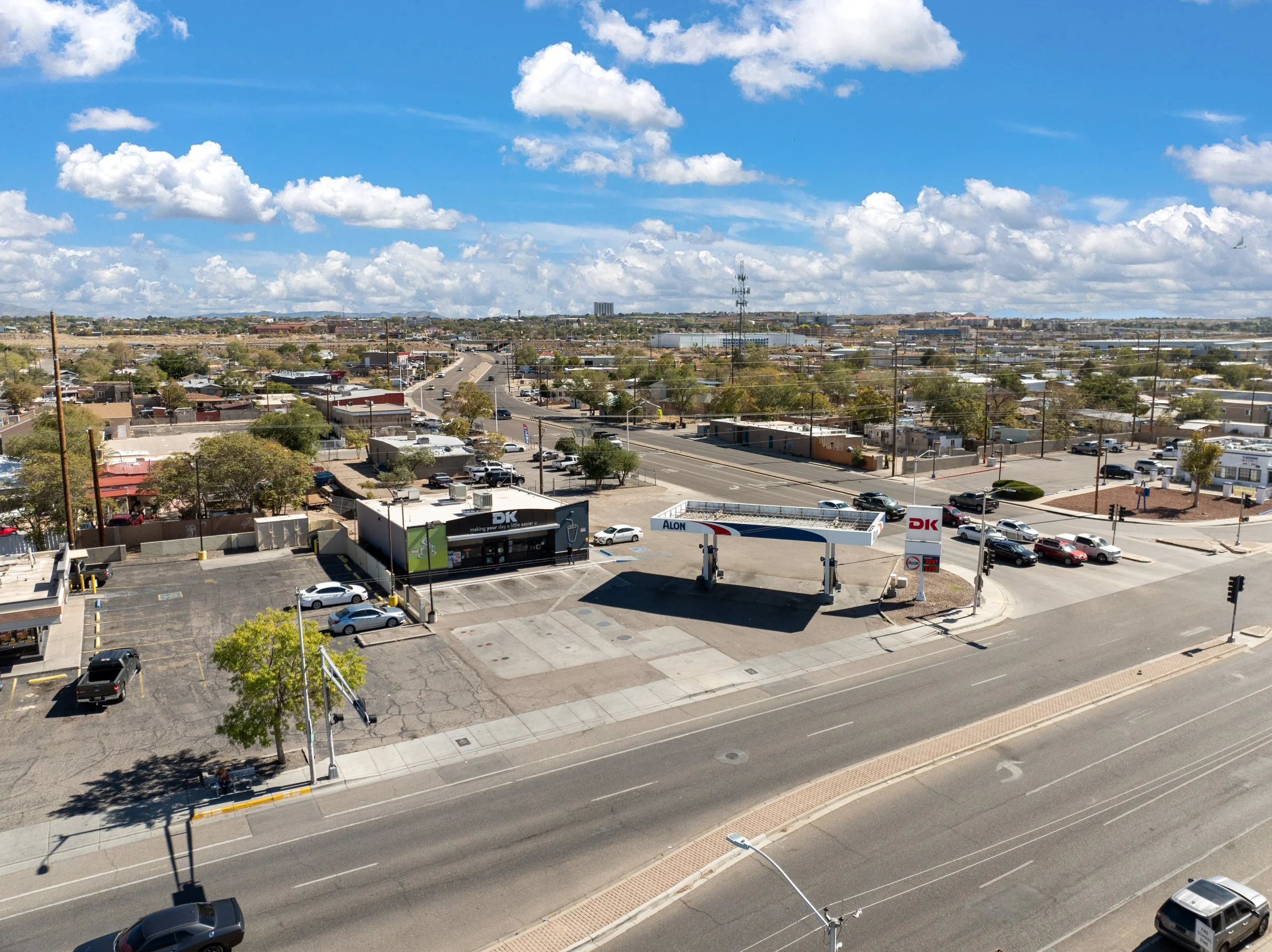 A cityscape with a clear blue sky filled with fluffy white clouds. The area includes commercial buildings, roads with vehicles, and a parking lot in the foreground. Power lines and a telephone tower are visible against the sky.