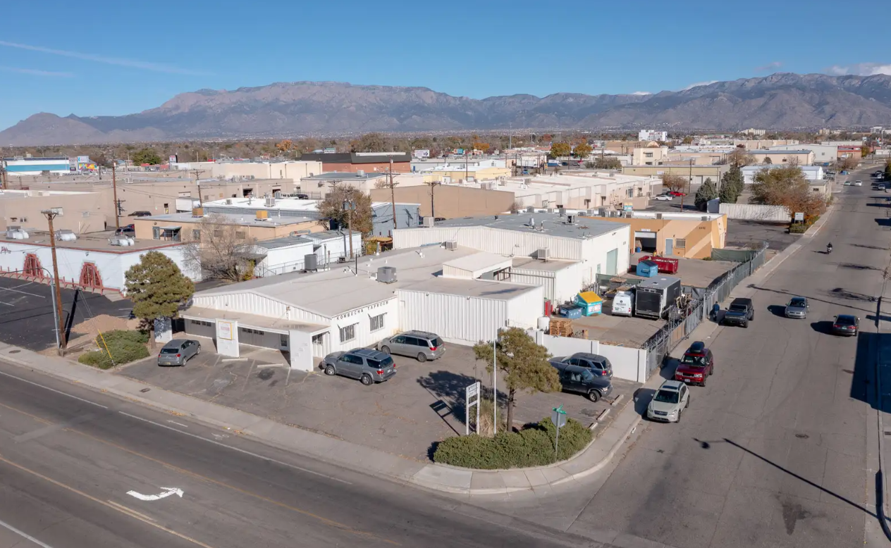 An aerial view of a commercial area with warehouses, parking lots, and a street with cars. Mountain range in the background under a clear blue sky.