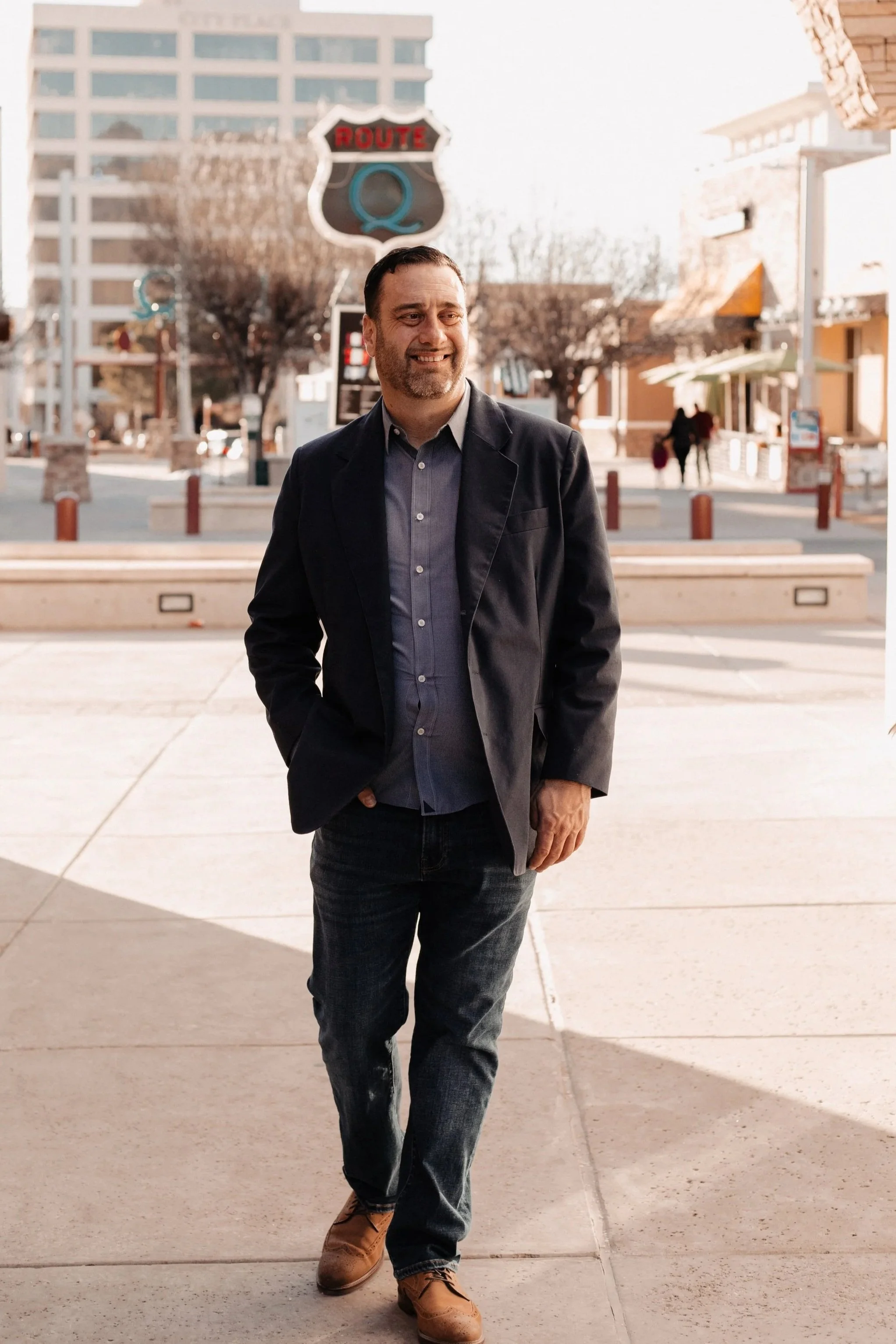 A man in a dark blazer, blue shirt, and jeans walking outdoors on a sunny day, with buildings and trees in the background.