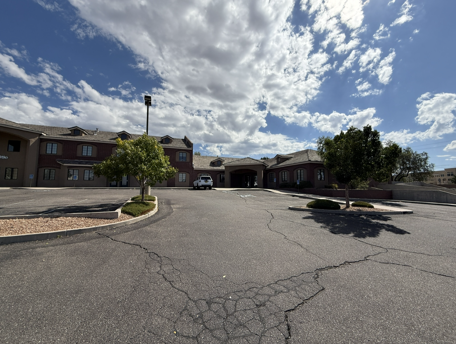 Empty parking lot in front of a building with trees and cloudy sky.
