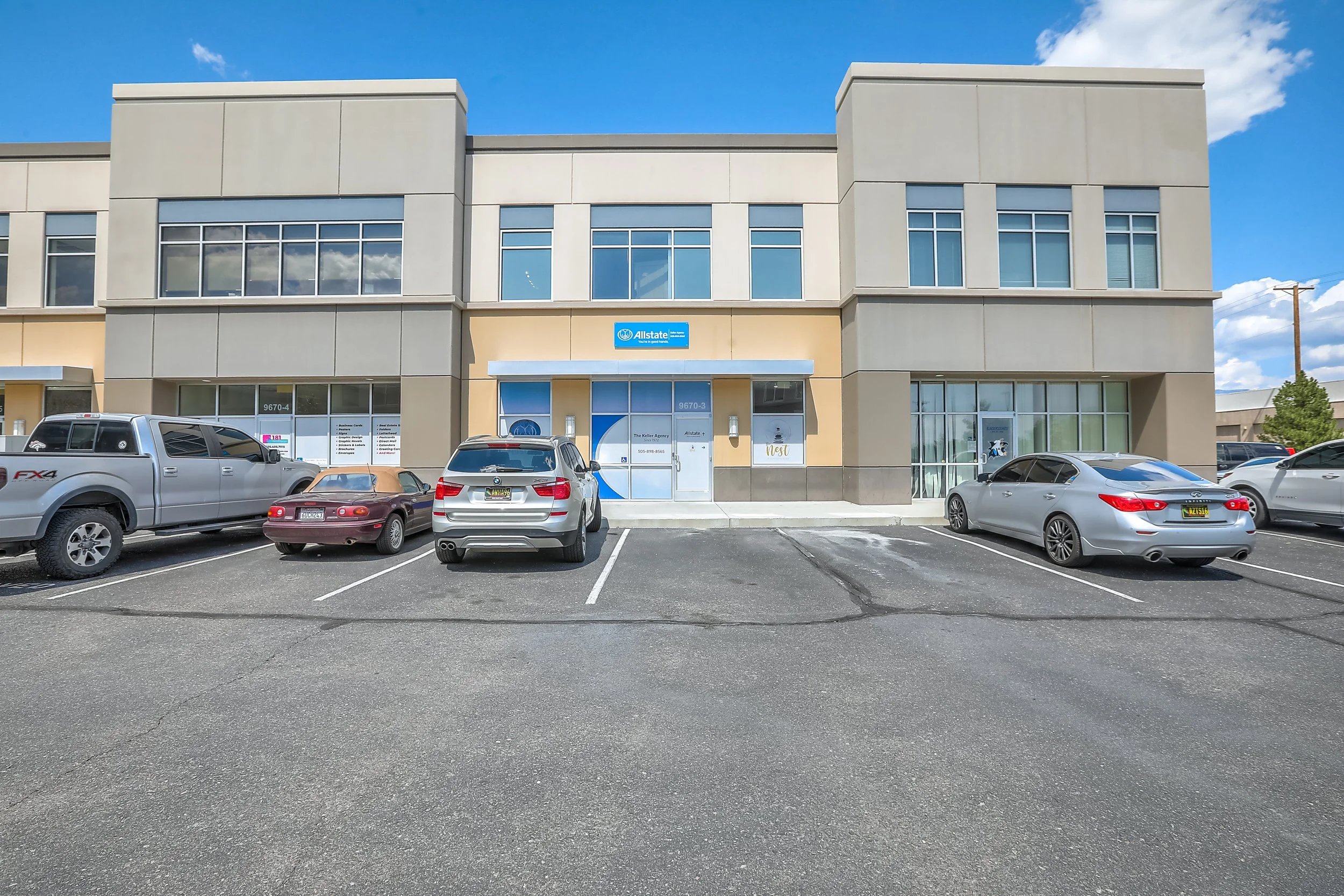 A beige commercial building with large windows and a blue Allstate logo sign in a parking lot with several parked cars, under a partly cloudy blue sky.