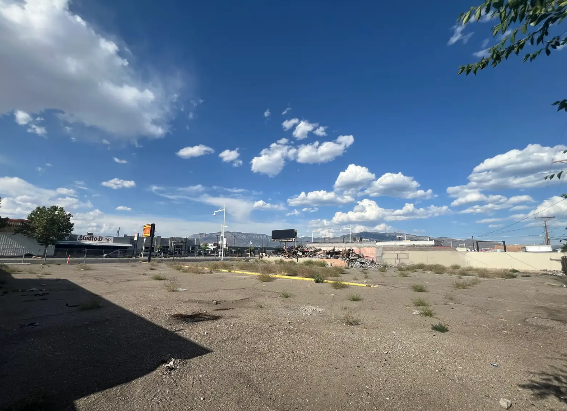 An empty parking lot with sparse weeds, a cloudy sky, and a view of distant mountains.