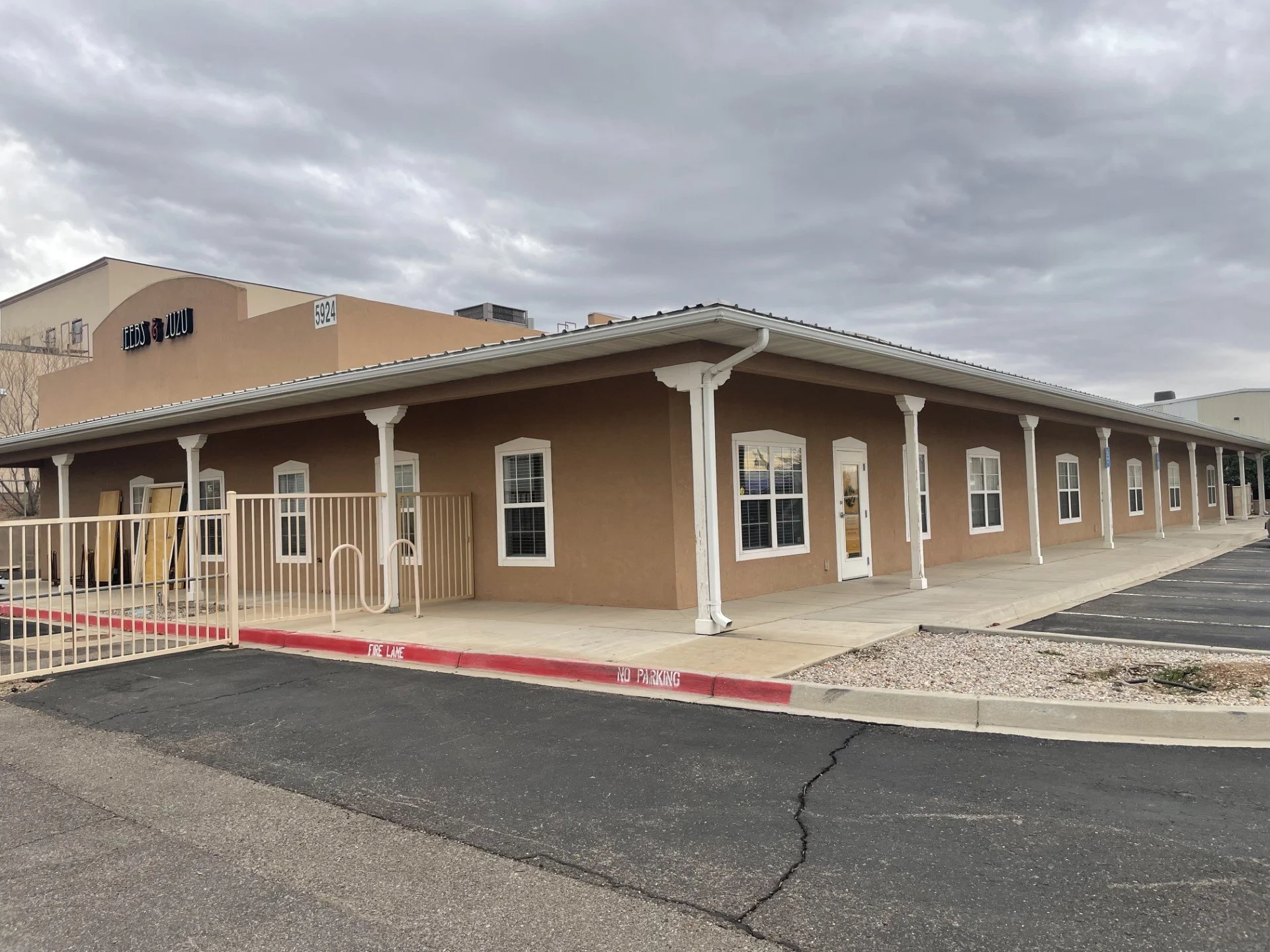 A beige building with white trim and a covered walkway with white columns. The building has multiple windows and a glass door. There is a sign with the text 'FERRAZ LUZU' on the upper part of the building. The parking lot in front has marked spaces and a red curb with the sign 'NO PARKING'. The sky is overcast.