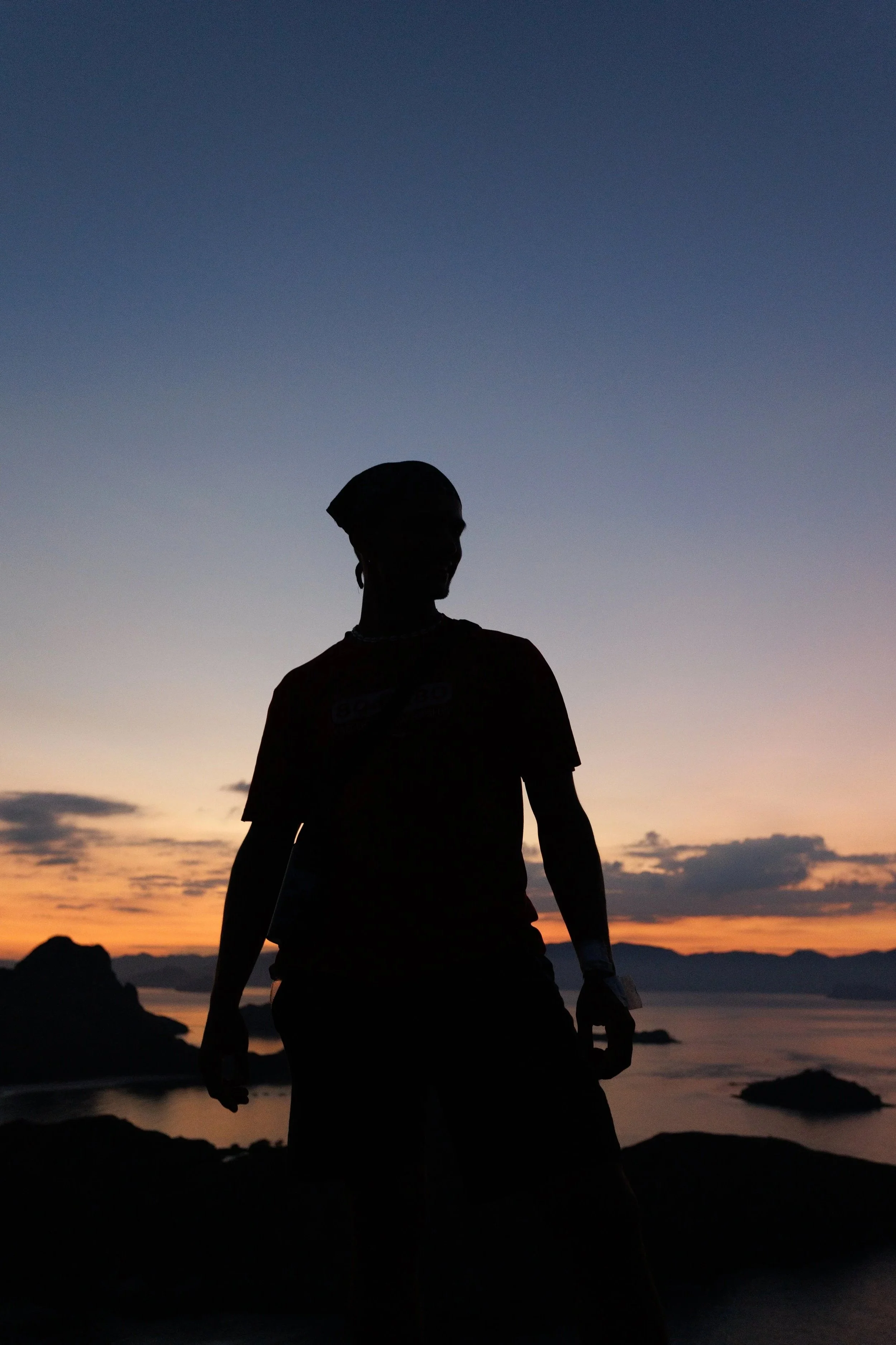Silhouette of a person standing outdoors at sunset with mountains and water in the background.