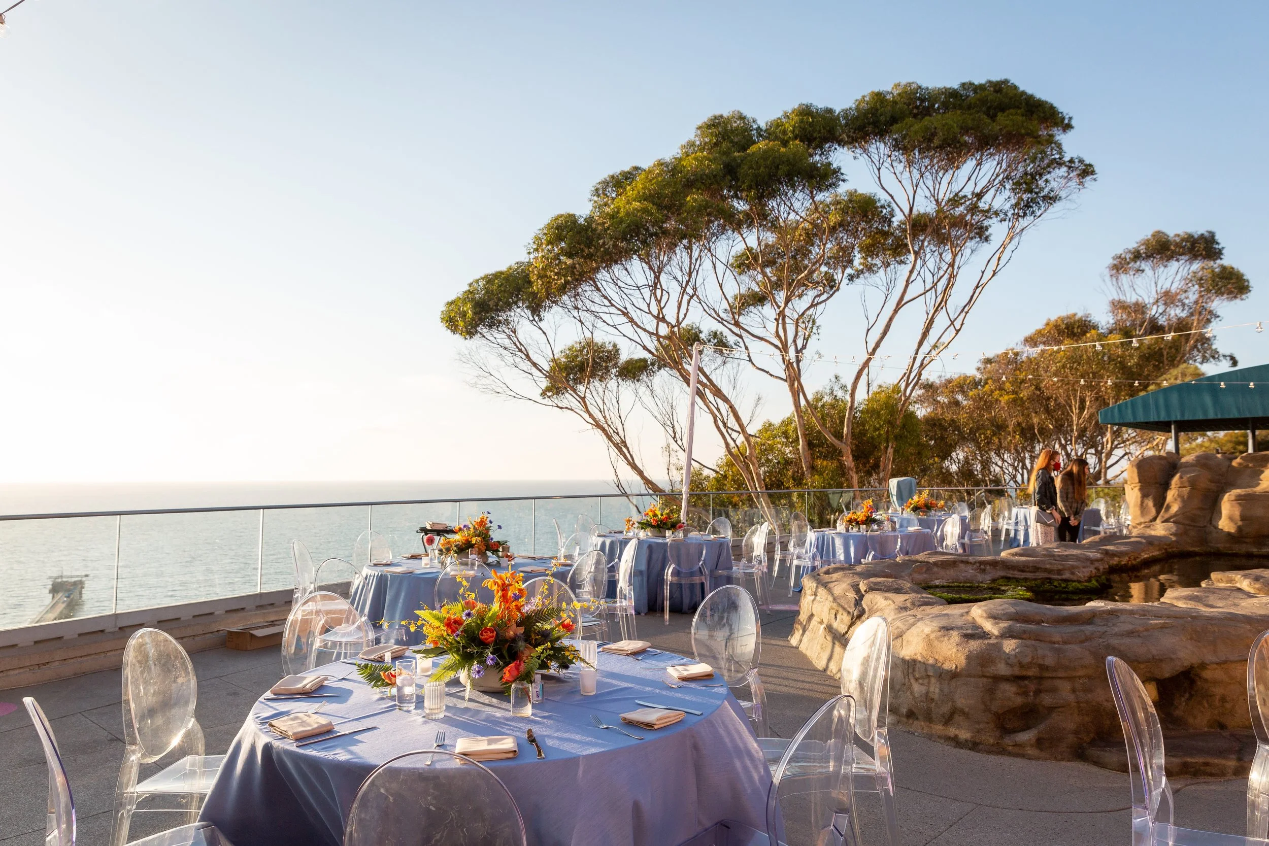Outdoor dining area with round tables covered in blue tablecloths, decorated with colorful floral centerpieces, clear chairs, overlooking the ocean with a glass railing, trees, and a rock formation in the background during sunset.