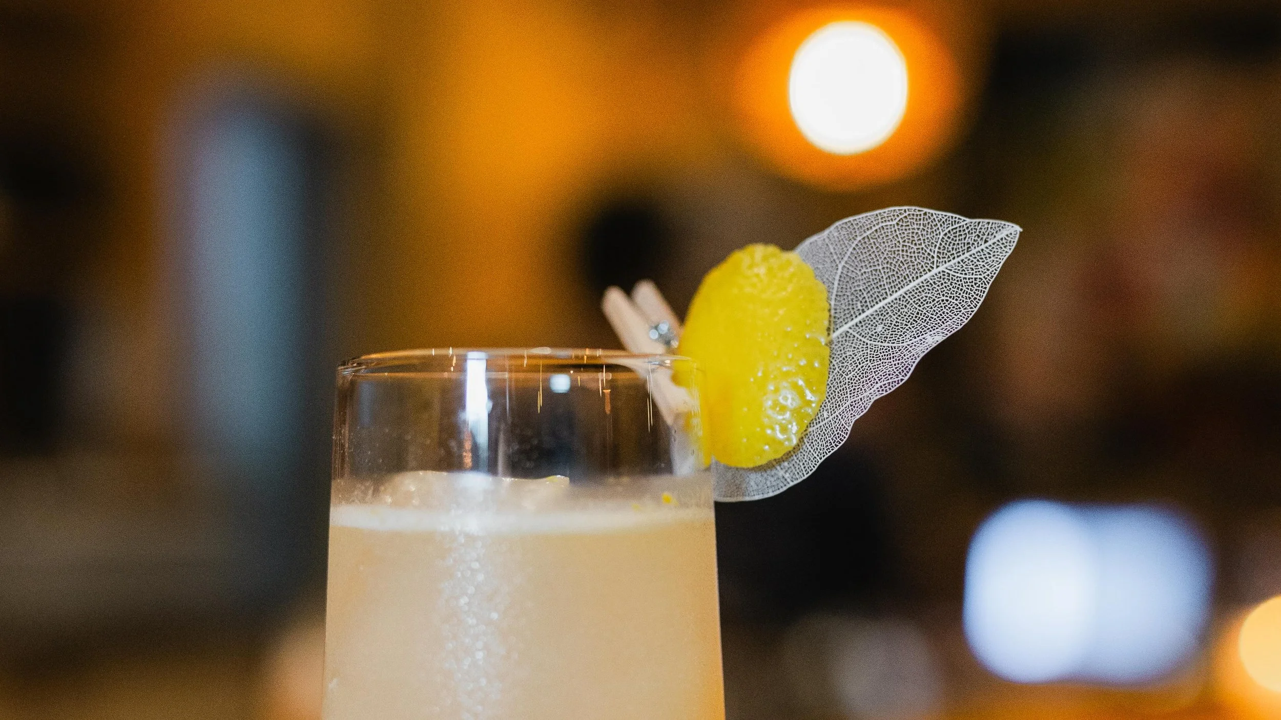 A close-up of a tropical cocktail garnished with a lemon wedge, a decorative leaf, and a small clothespin, with a warm, blurred background.