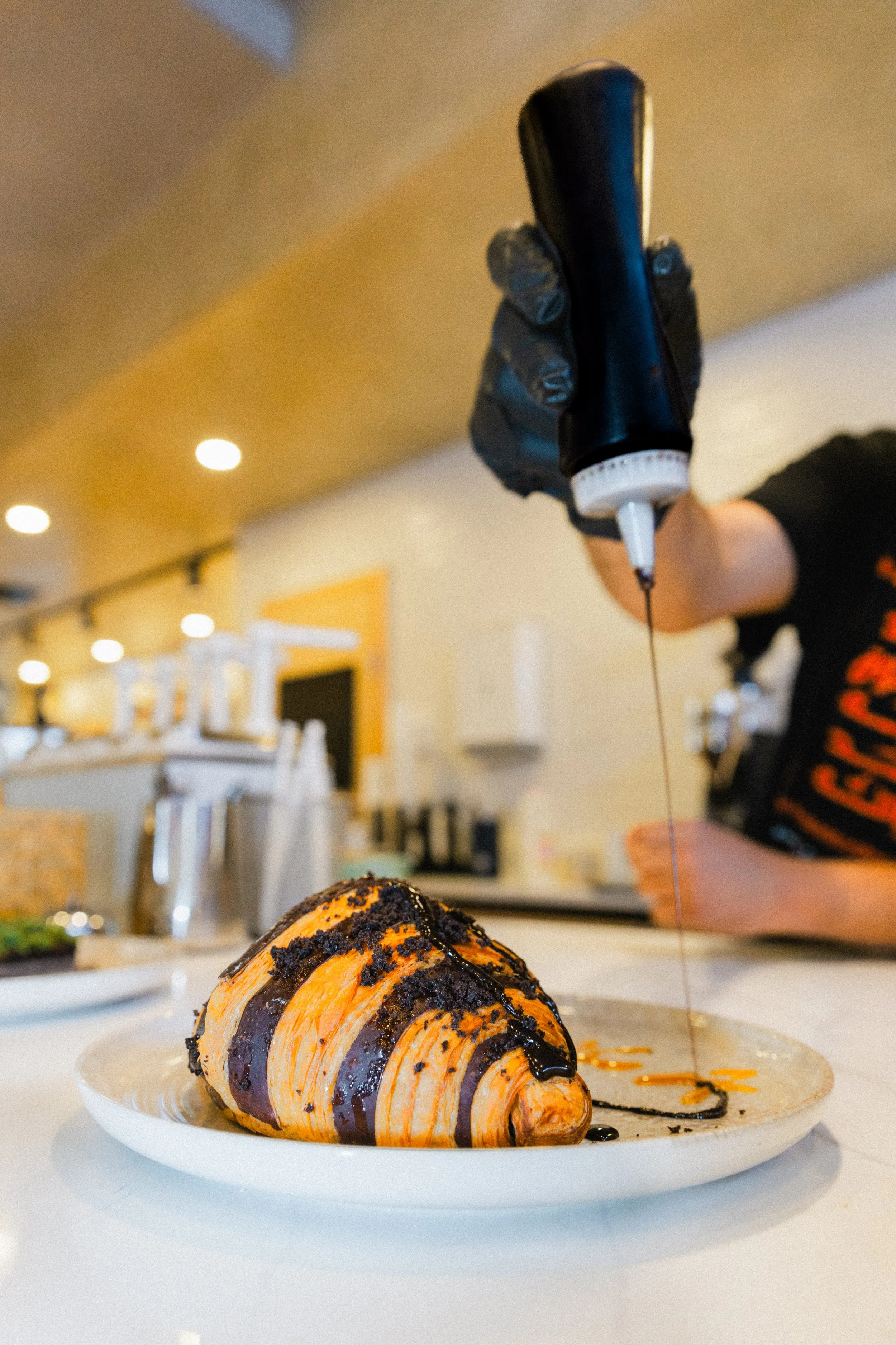 A chef decorates a croissant with chocolate syrup from a squeeze bottle.