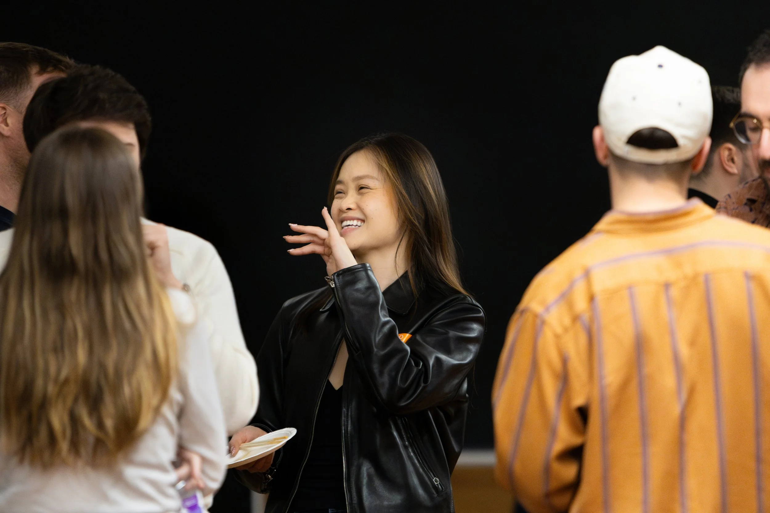 A group of diverse young adults socializing, with a woman in a black leather jacket smiling and touching her face.