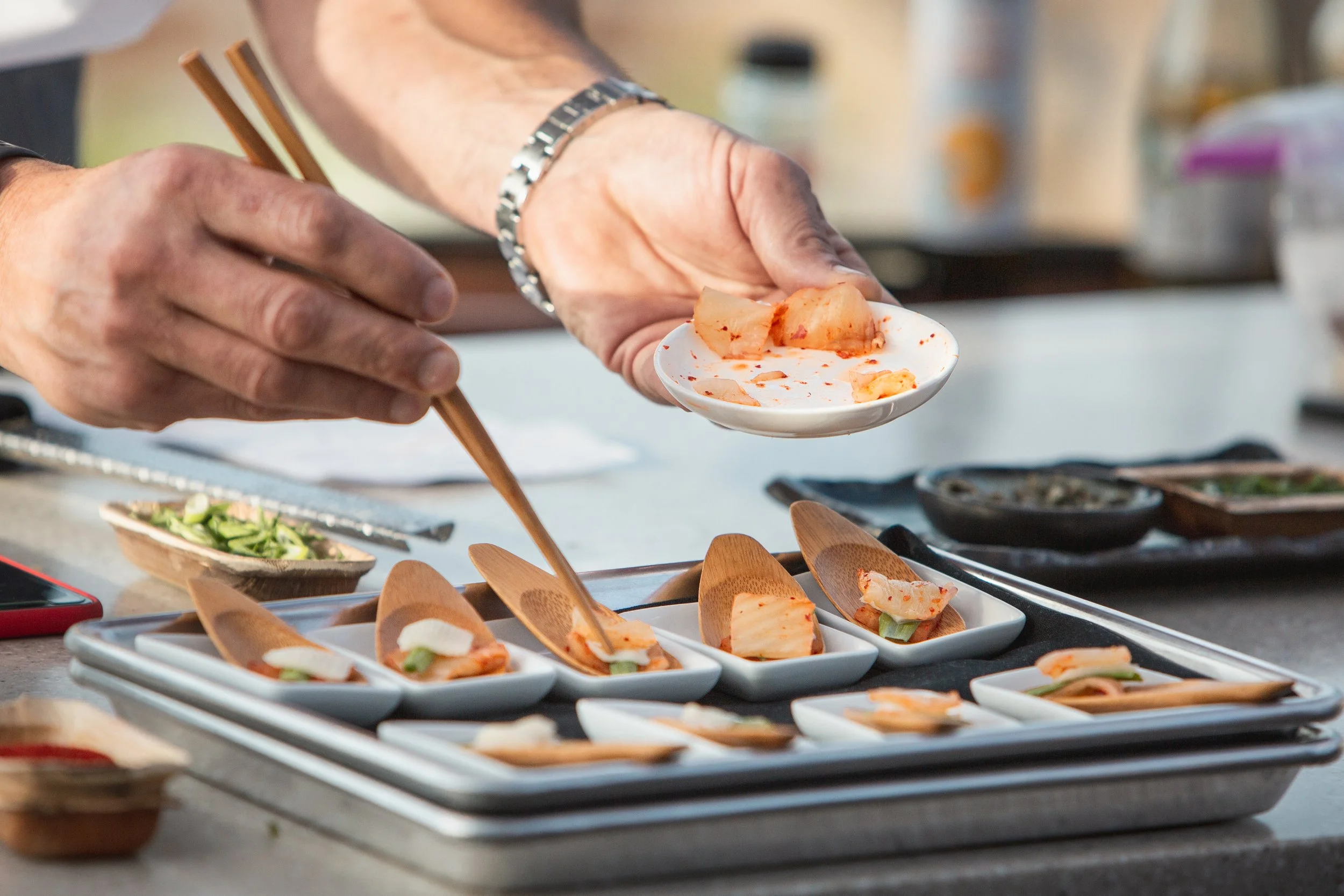 Person using chopsticks to place kimchi on small white plates, with various kimchi servings on the table.