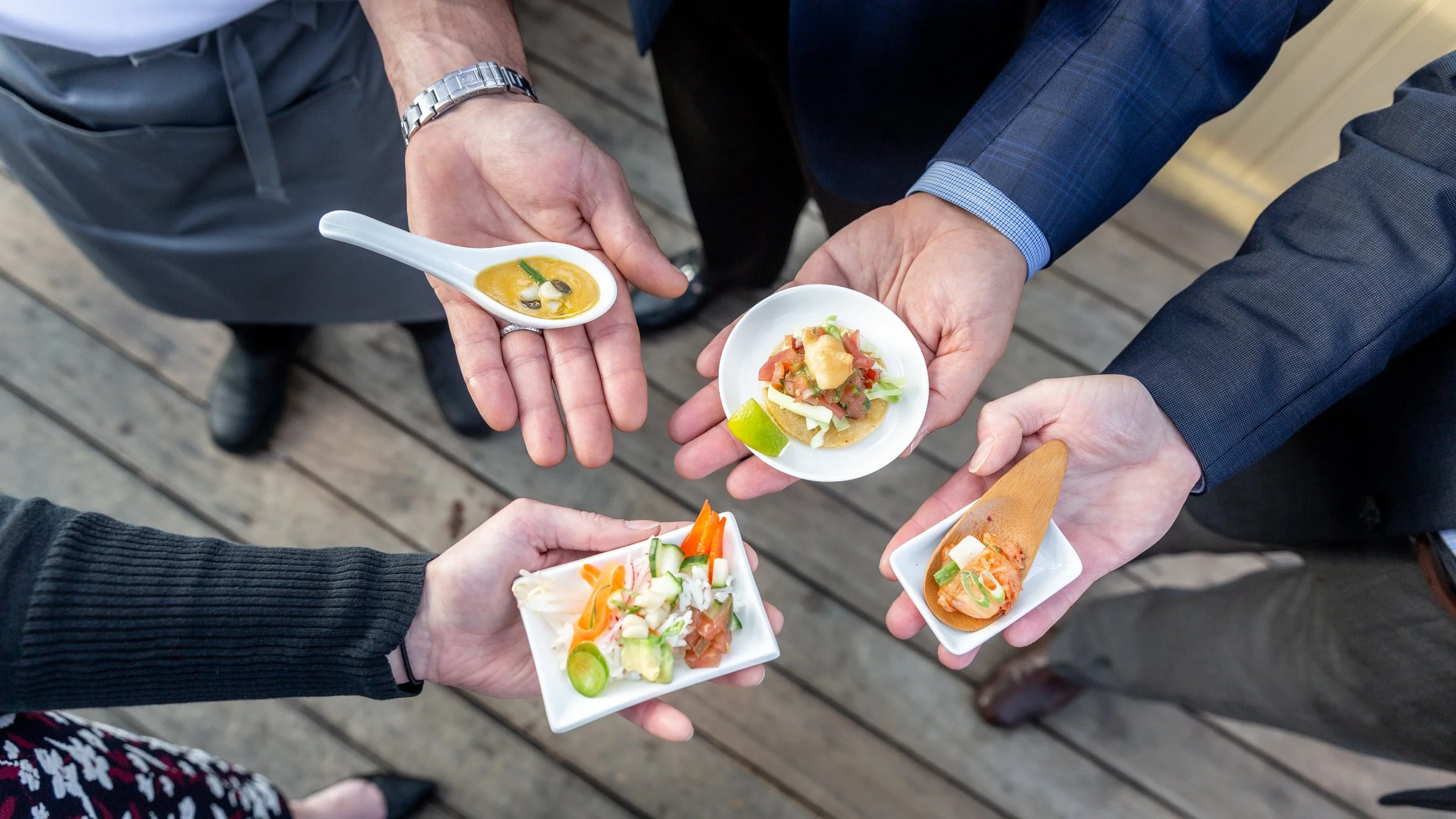 People holding various small plates of food including soup, salad, guacamole, and ceviche on a wooden deck.