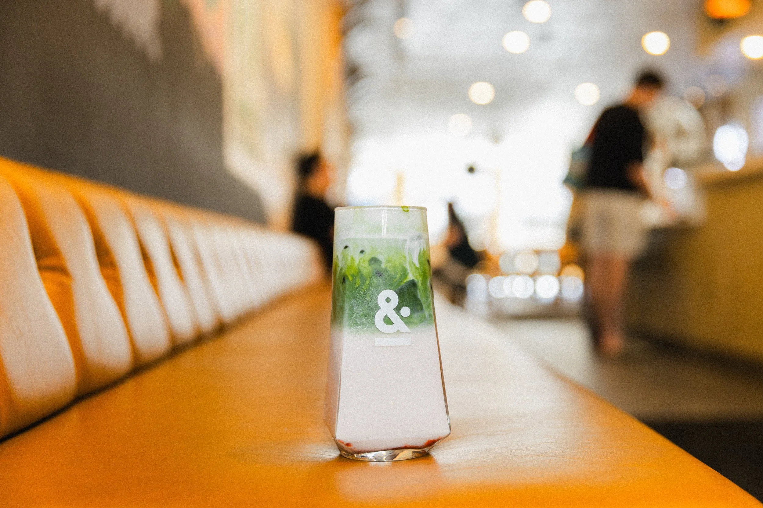 A glass with a layered drink, featuring green, white, and red colors, placed on an orange table in a cafe. The background shows blurred people and cafe interior