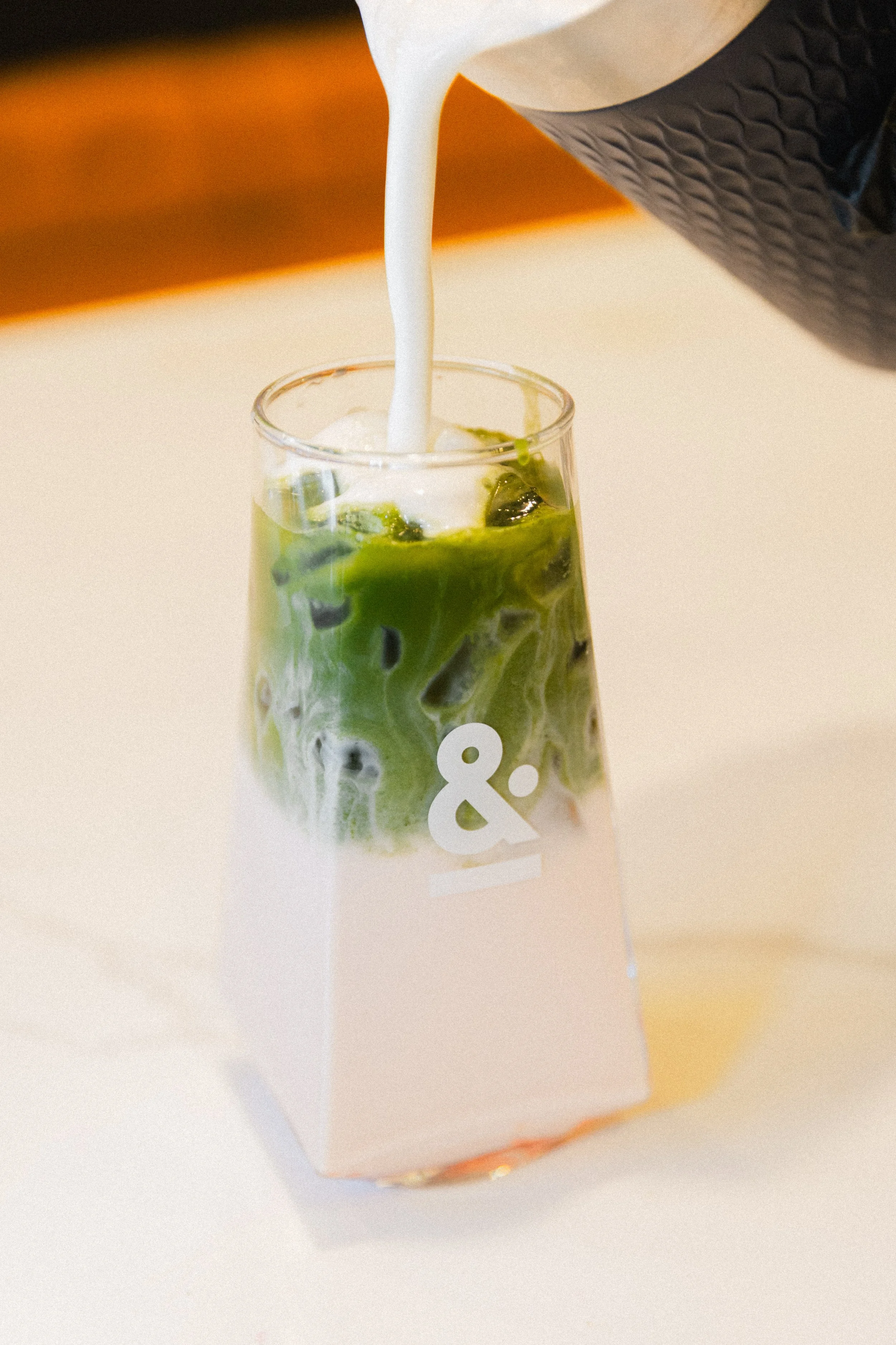 A tall glass of iced matcha latte with green tea and milk, with milk being poured into it.